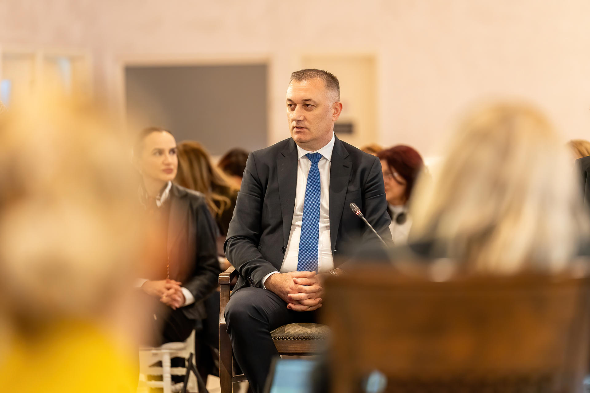 Photo of a formal event; man in a dark suit and blue tie seated among attendees.