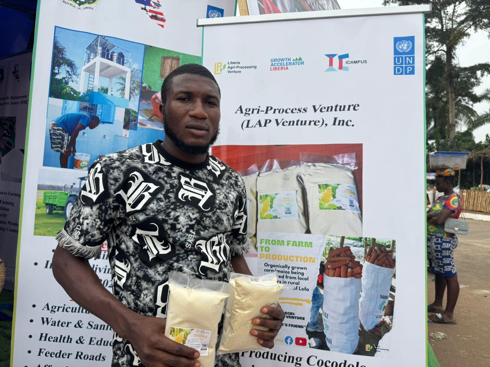 Person in a black-and-white patterned shirt holds a booklet in front of an agricultural exhibition display board.