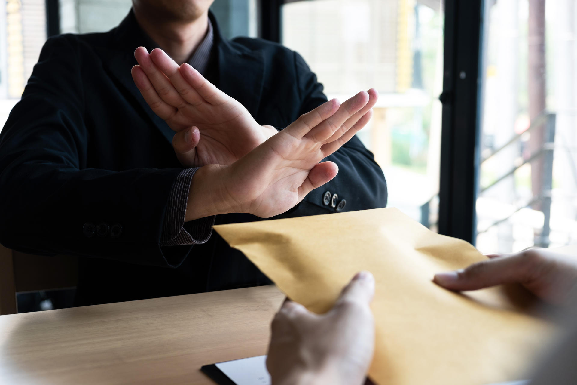Person in a dark suit raises hands in a stop gesture across a desk; envelope on the table.