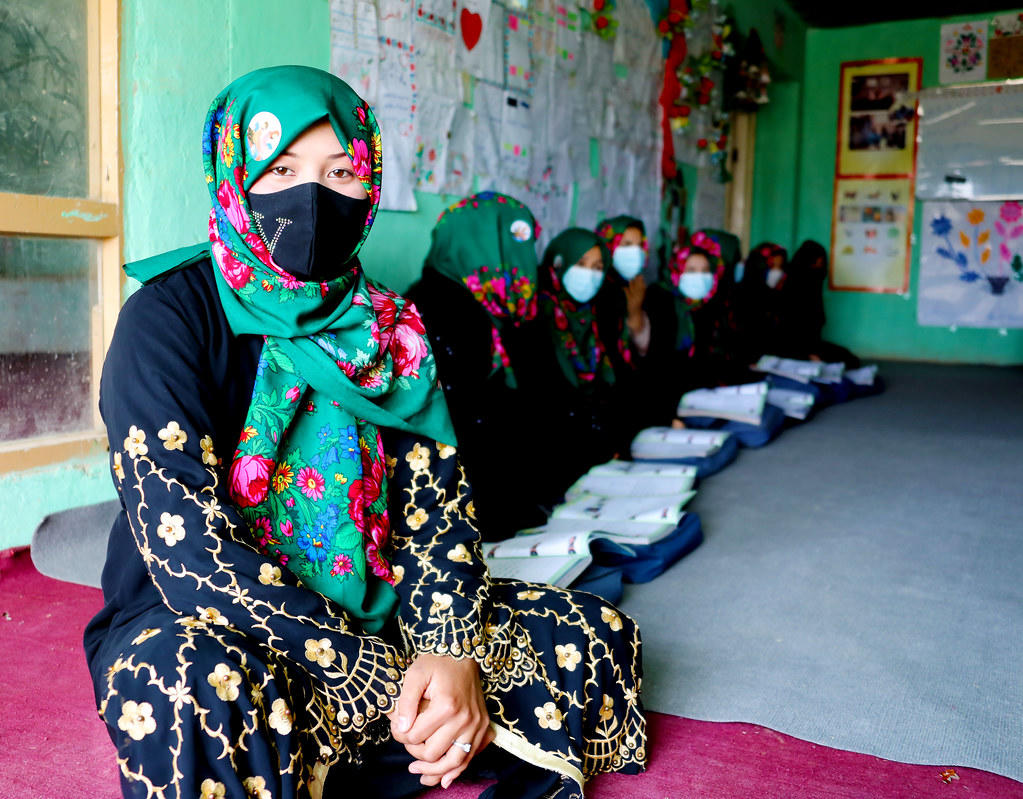 Group of women in colorful hijabs and masks sitting on mats in a classroom; foreground woman in teal hijab.