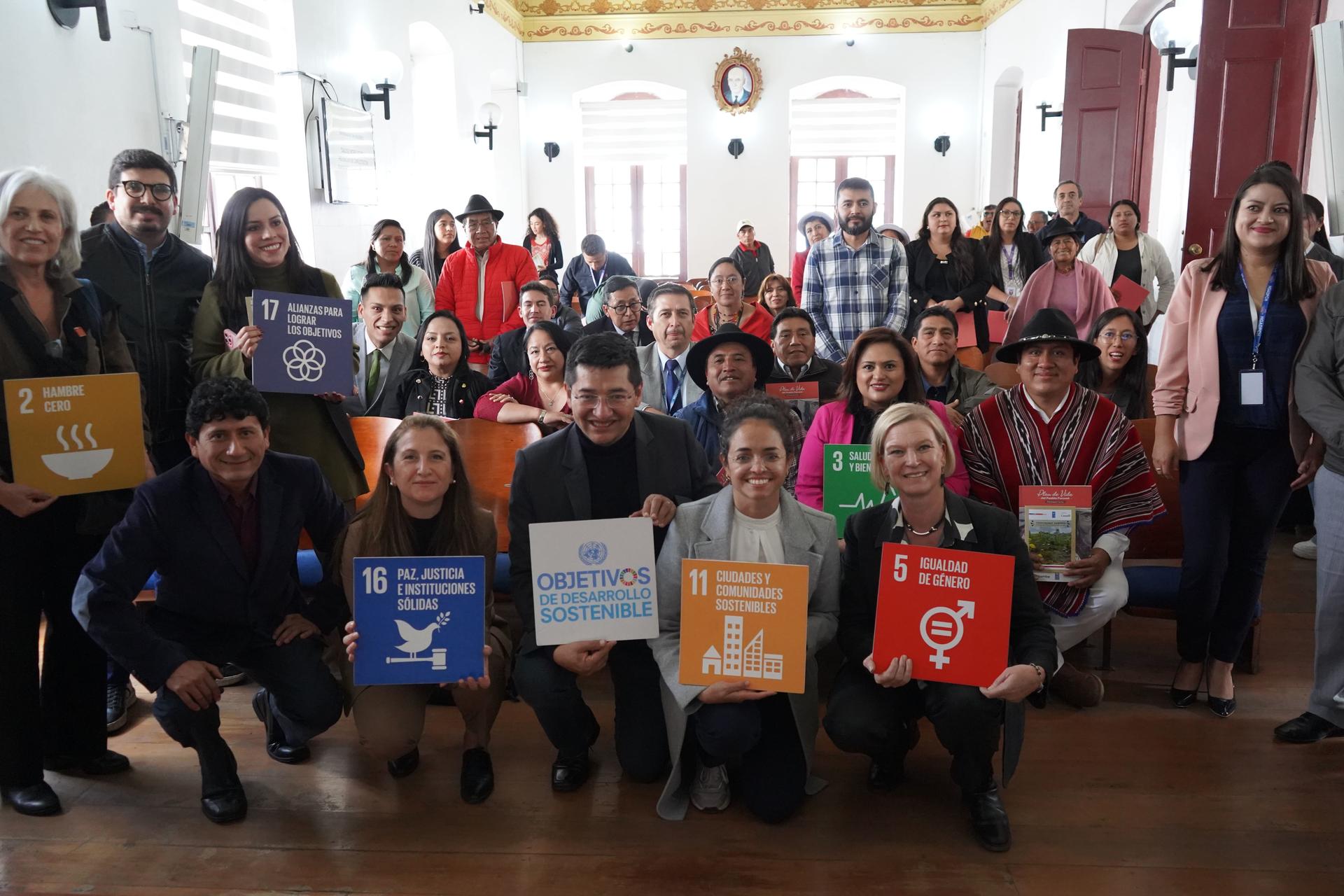 Group photo: diverse people in a bright room, standing and kneeling, holding colorful signs.