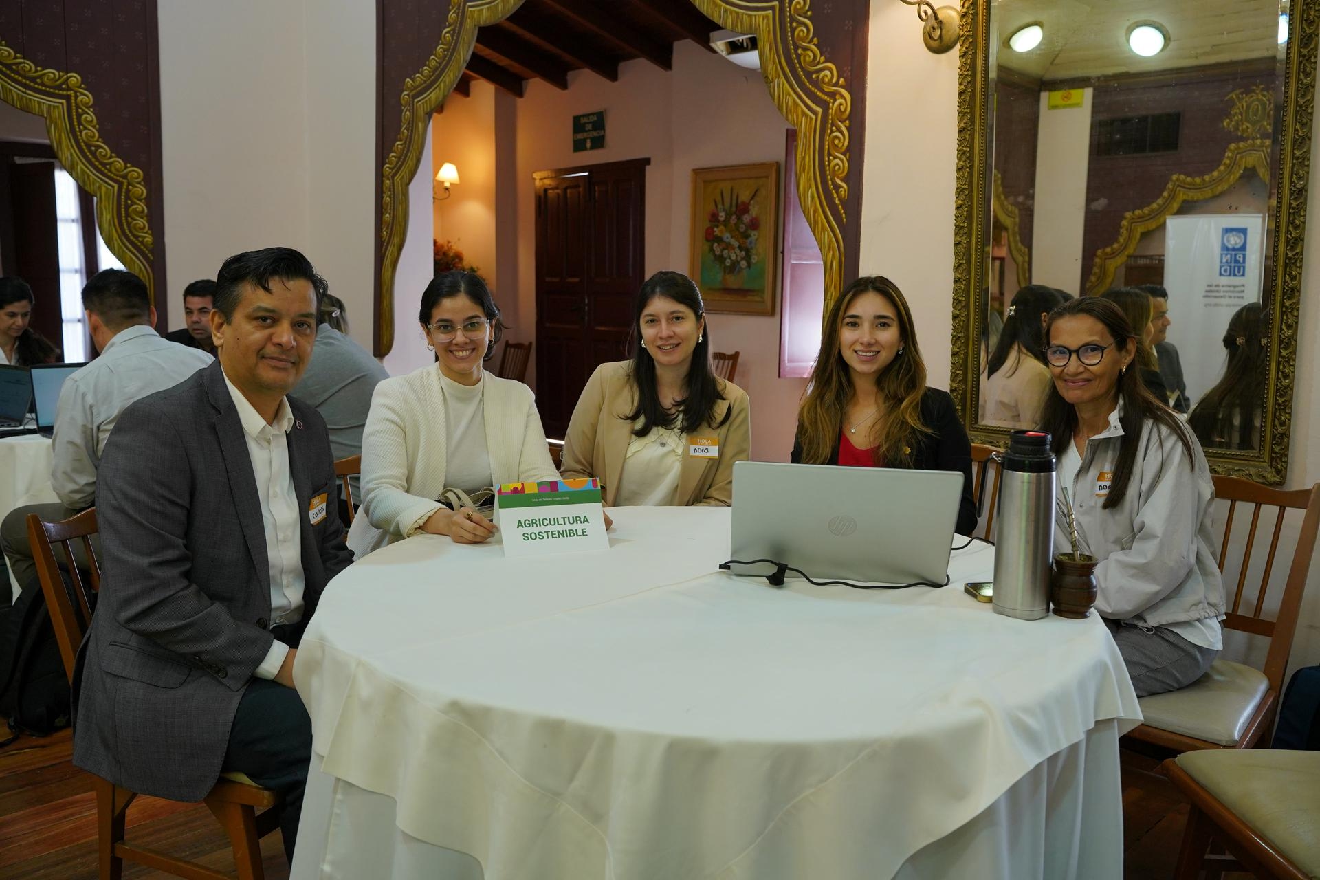 Five people pose around a round table with a laptop, smiling in an ornate, warmly lit room.