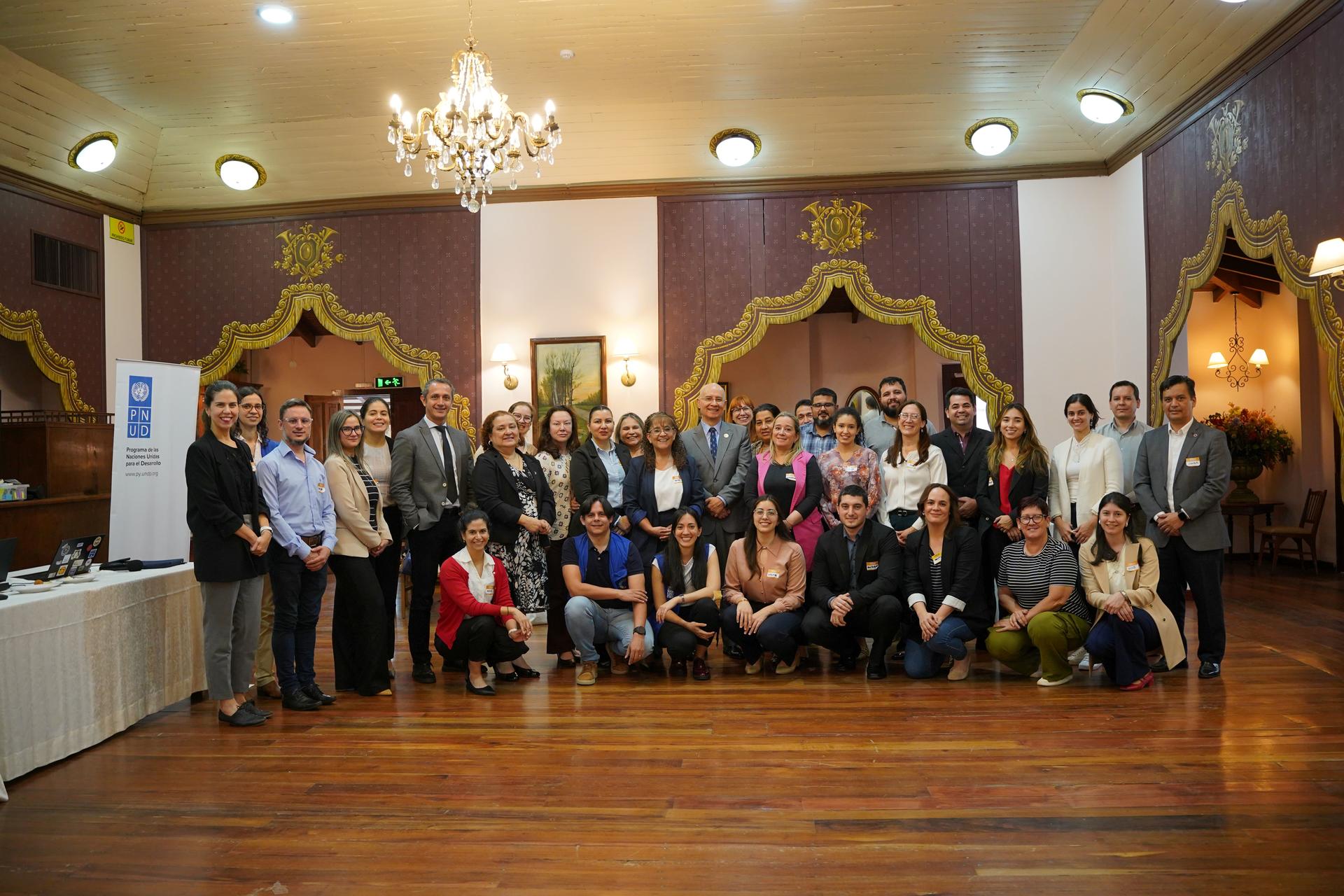 Group of adults posing for a formal photo in a grand hall with chandeliers.
