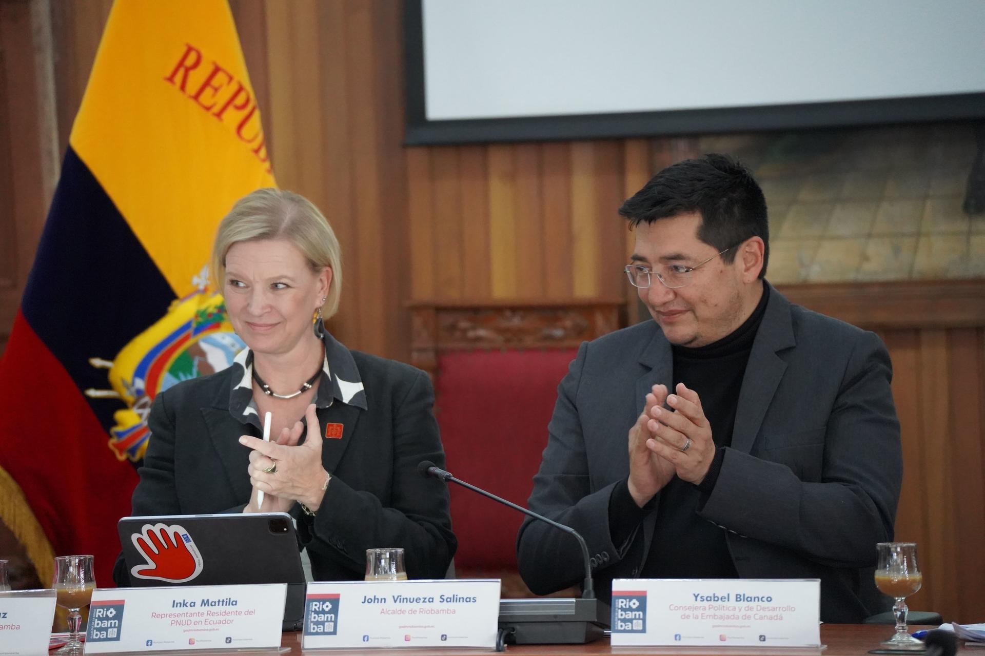 Two formally dressed officials sit at a table with nameplates and microphones, flags in background.