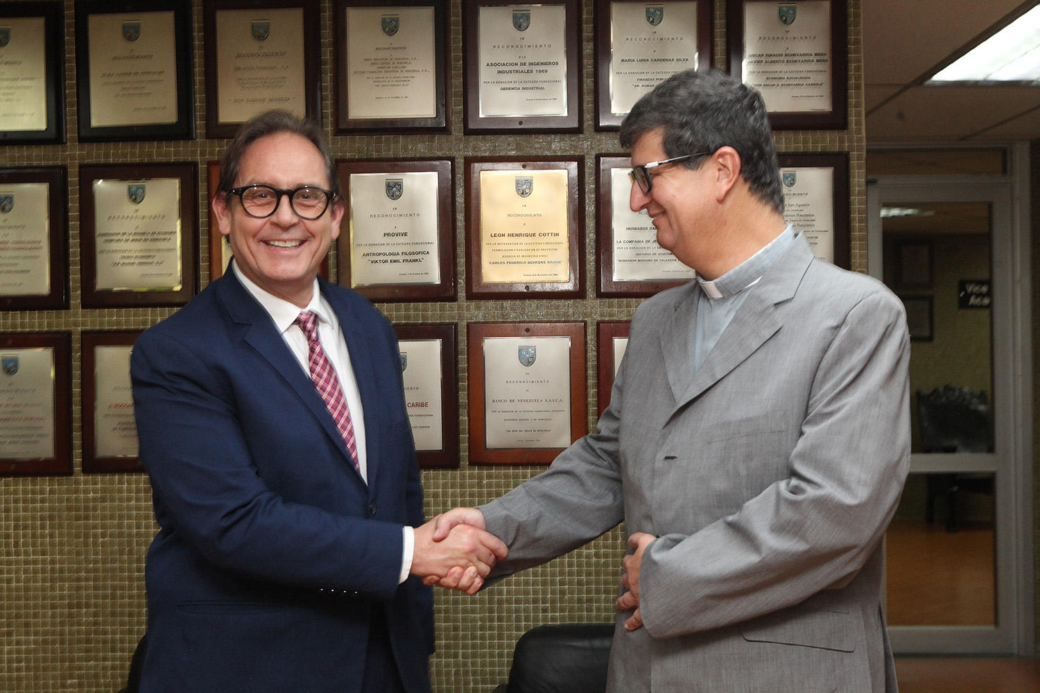 Two men in suits shake hands in front of a wall of framed certificates.