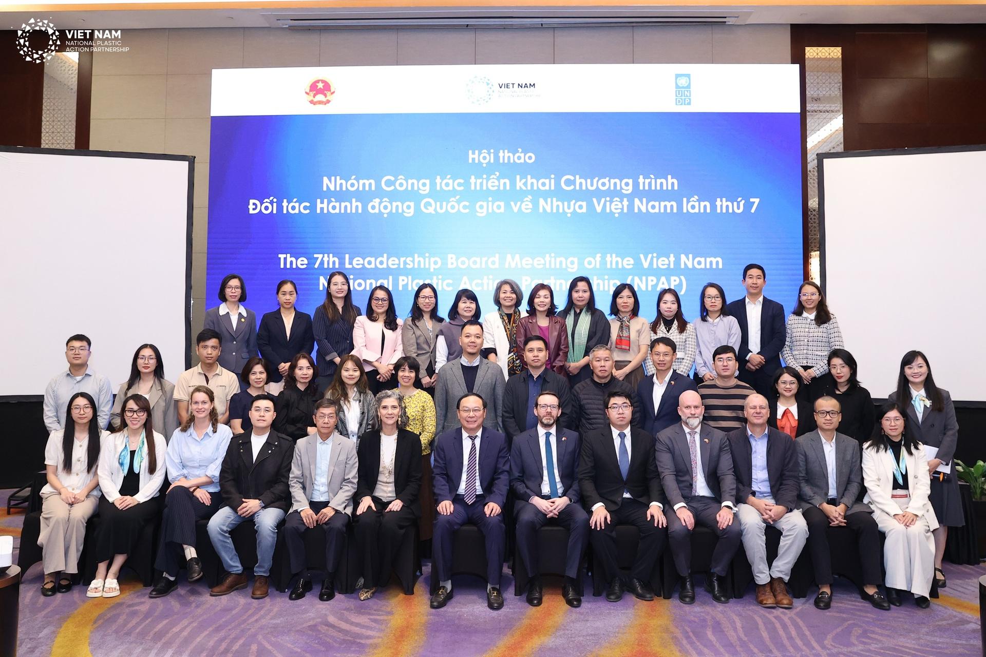 Group of formally dressed people posing on stage at a conference, banner with logos in background.