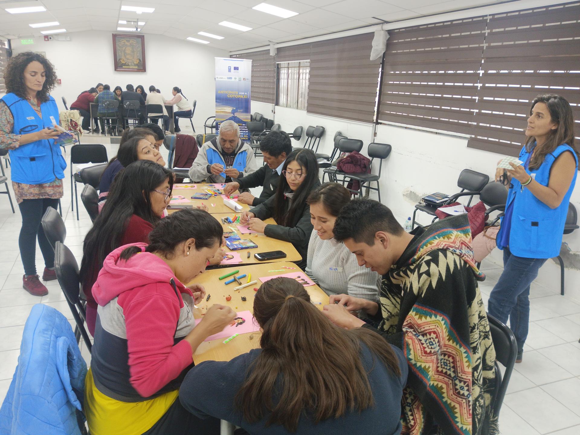 Group of people seated around tables in a bright classroom; two facilitators in blue stand nearby.