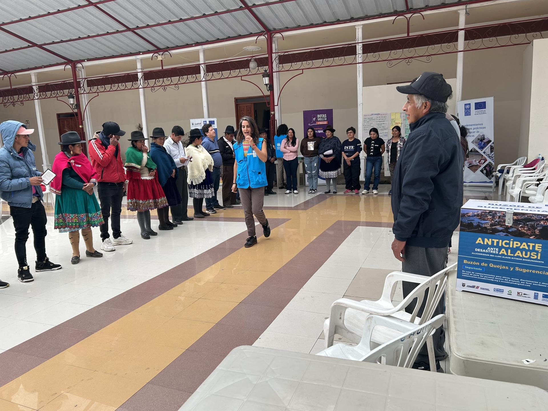Photograph of a man in a hat addressing a line of people in a hall with tables and banners.