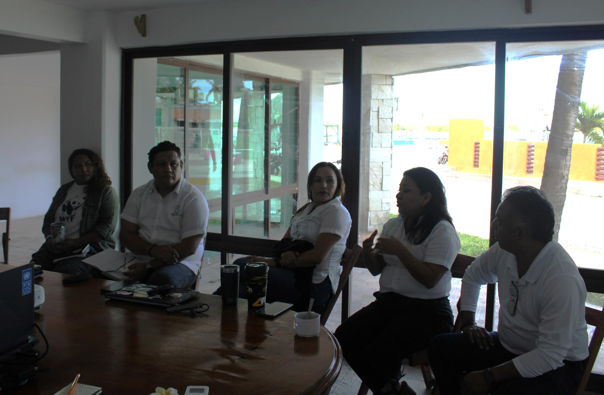Six people sit on the floor around a low table in a bright room with large windows.