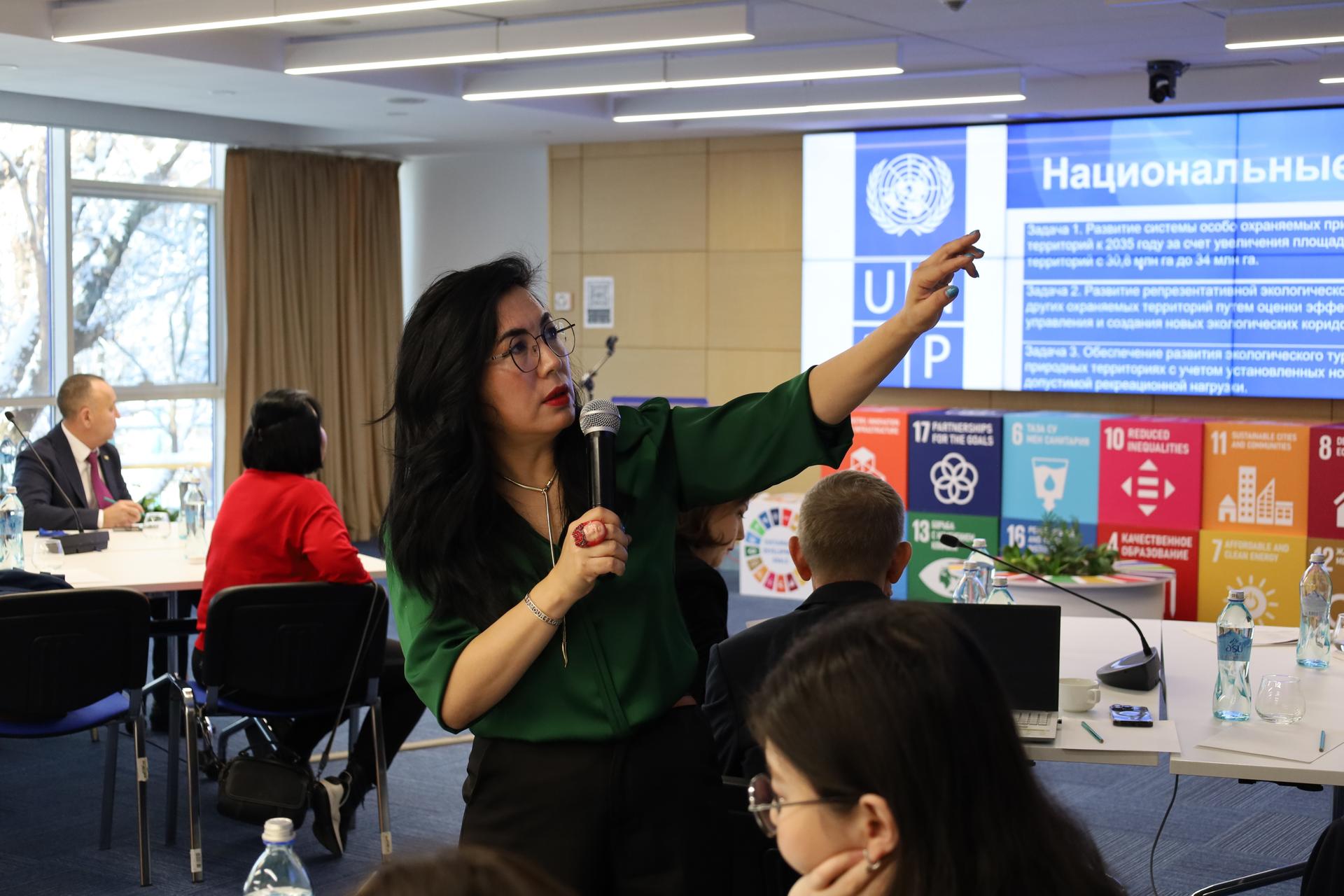 Female presenter speaks into a microphone in a conference room, pointing at a display.