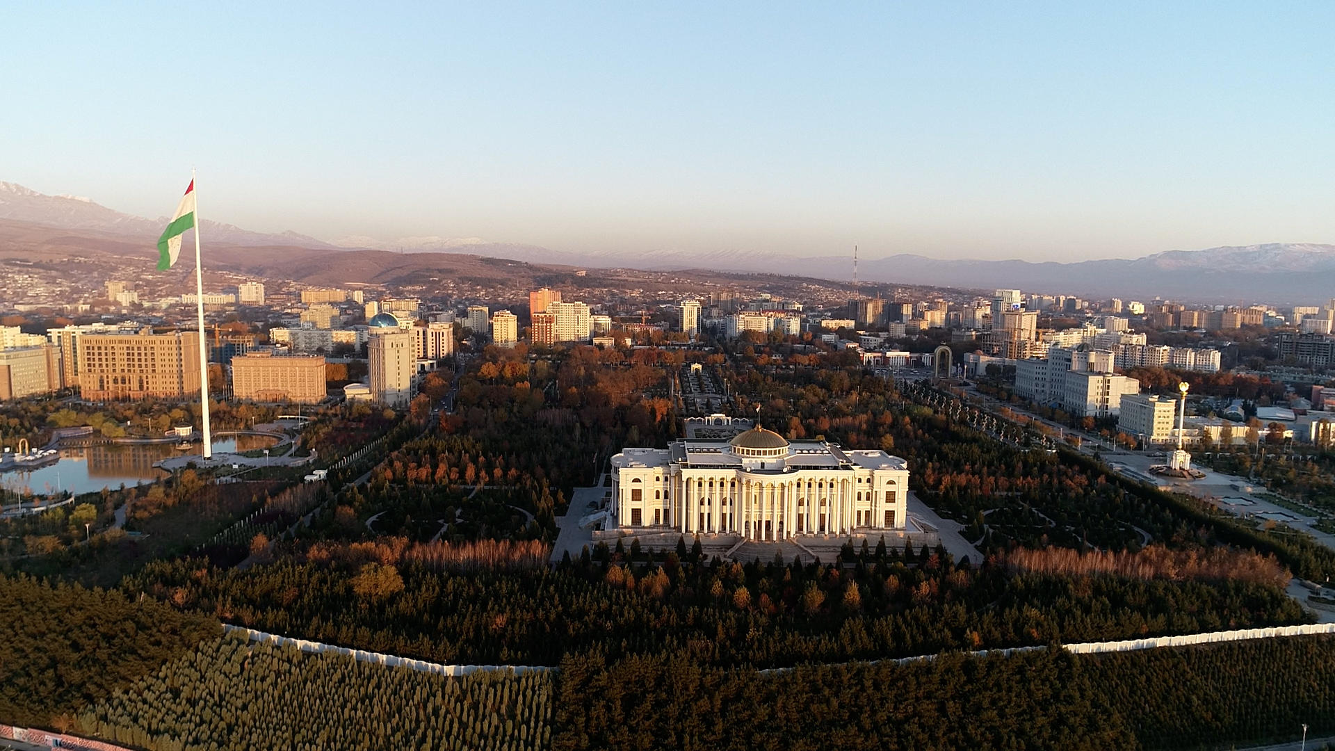 Aerial view of a city with a central white building, expansive green lawns, and distant hills.