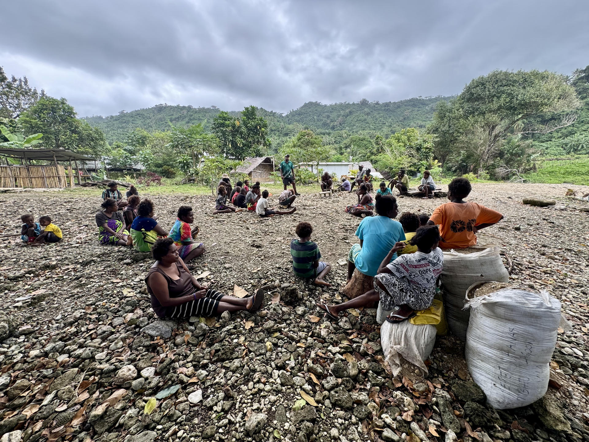 Photograph: rural riverbank gathering; people sit in groups, bags nearby, hills in distance.