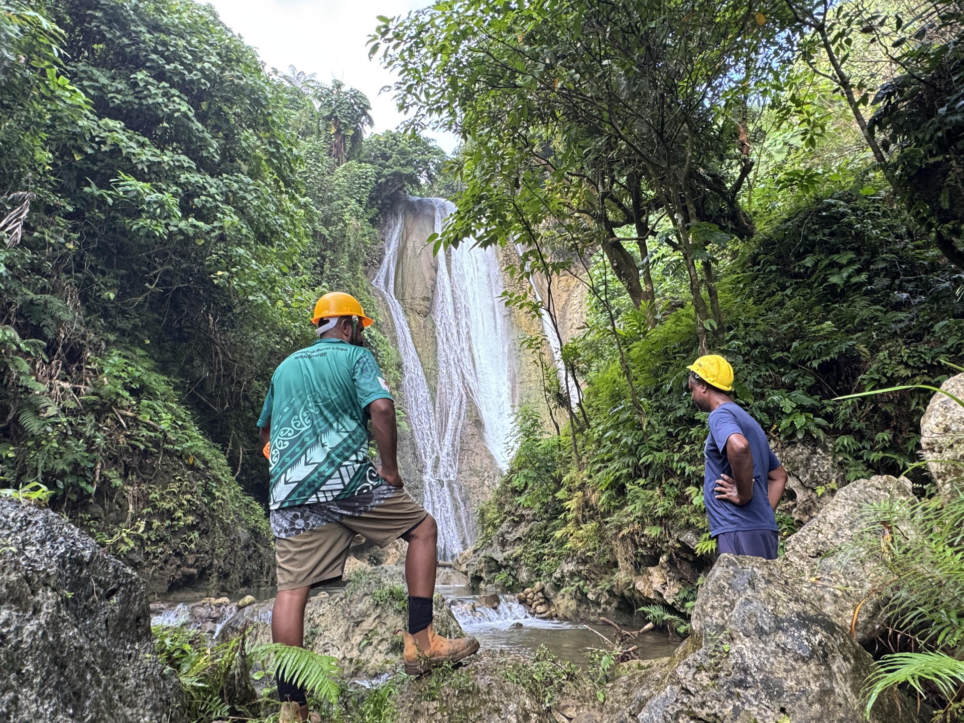 Two hikers wearing yellow helmets stand on rocks beside a tall waterfall in a lush forest.