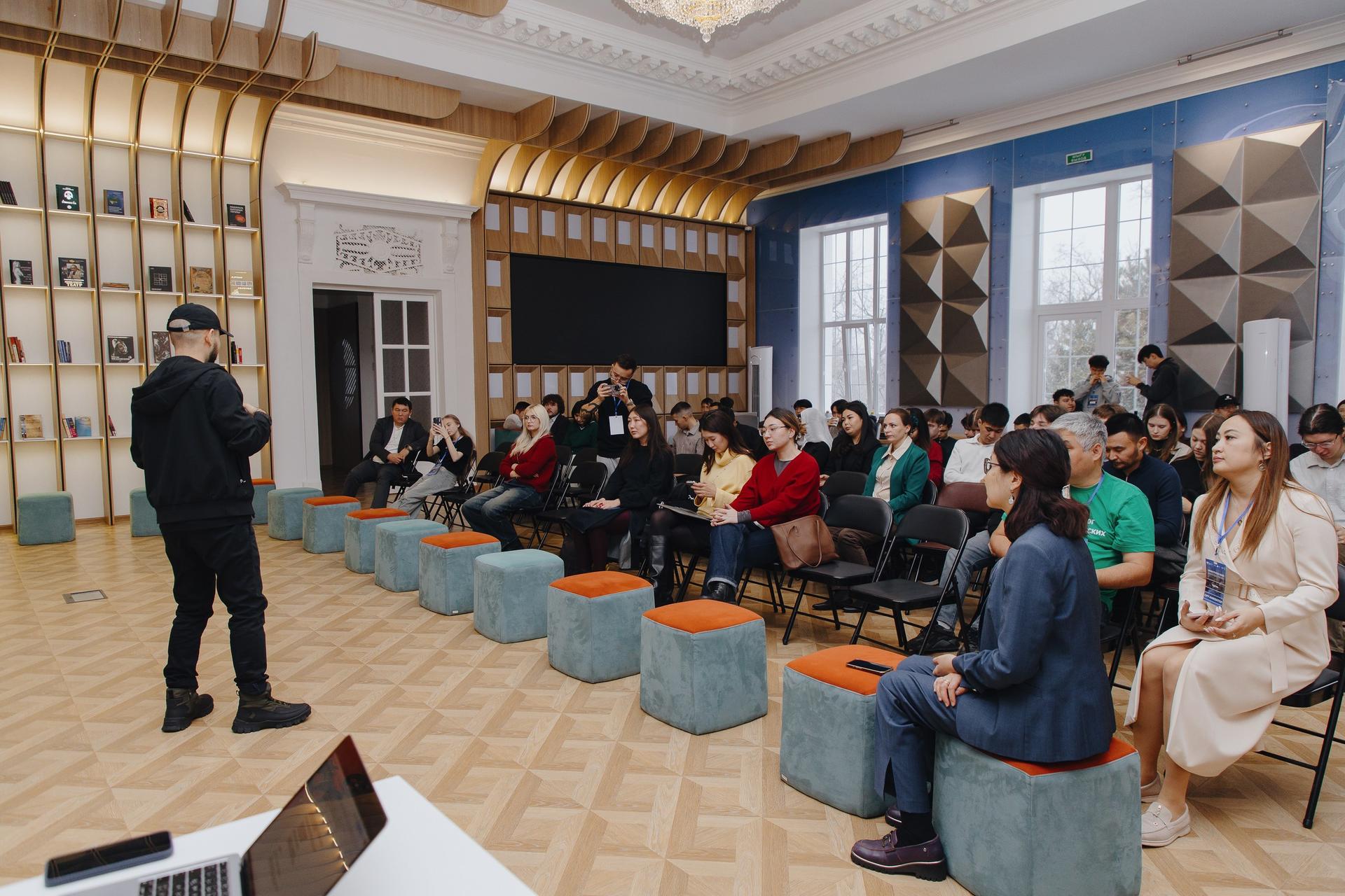 Presenter addresses a seated audience in a modern lecture room with colorful ottomans.
