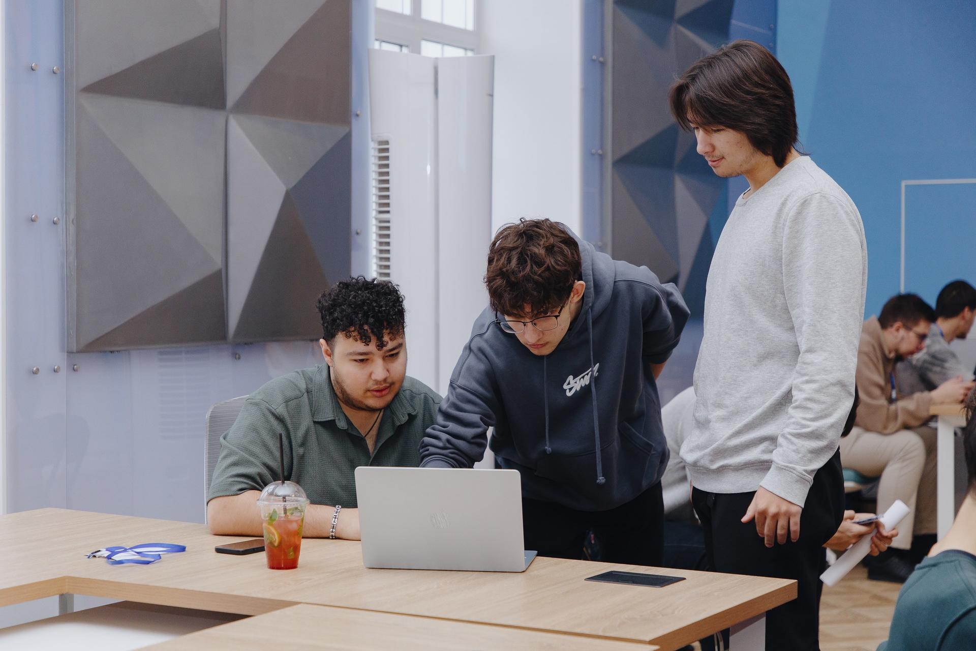 Three colleagues consult a laptop at a light wood table in a bright modern office.