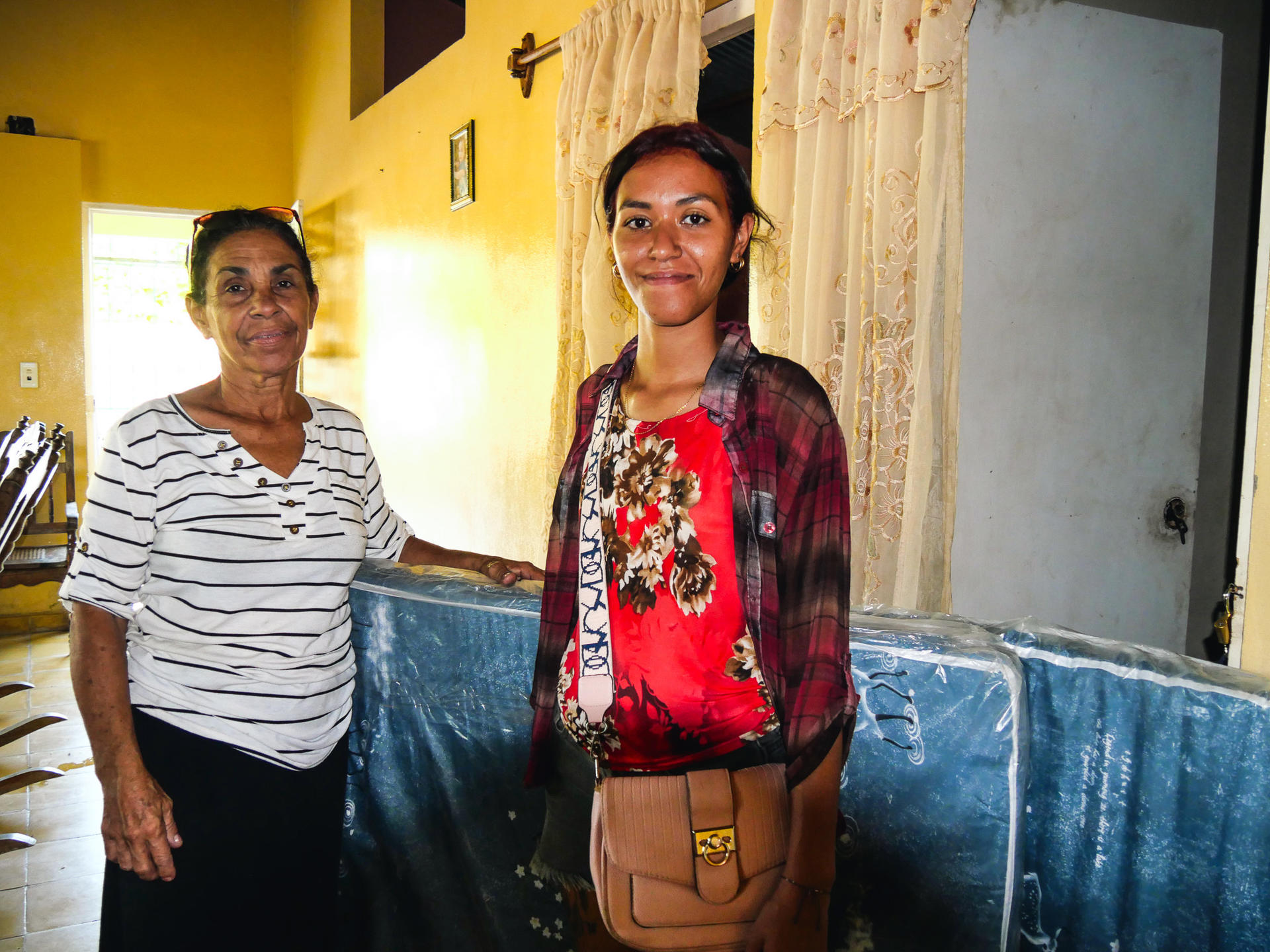 Two women stand in a yellow hallway; striped top and red floral blouse with tan bag.