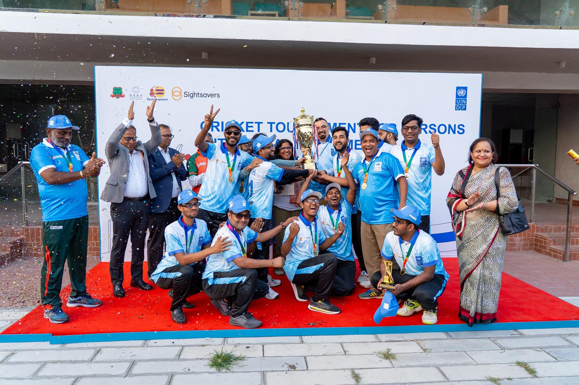 Group of people in blue shirts posing on a red carpet in front of a banner, some raising hands.