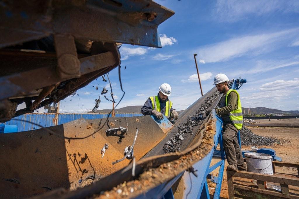 Two workers in hard hats and high-visibility vests supervise dumping from a blue industrial truck.
