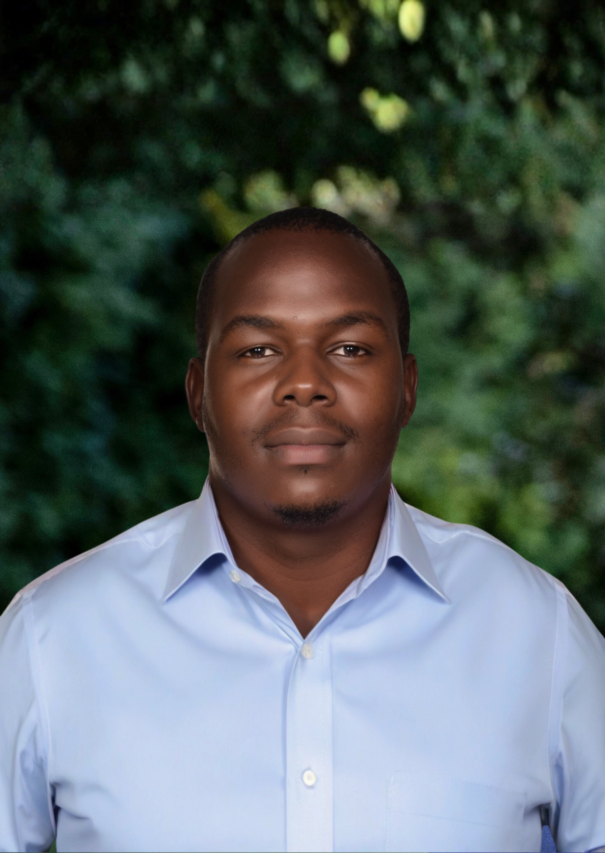 Man wearing a light blue shirt standing outdoors with green foliage behind.