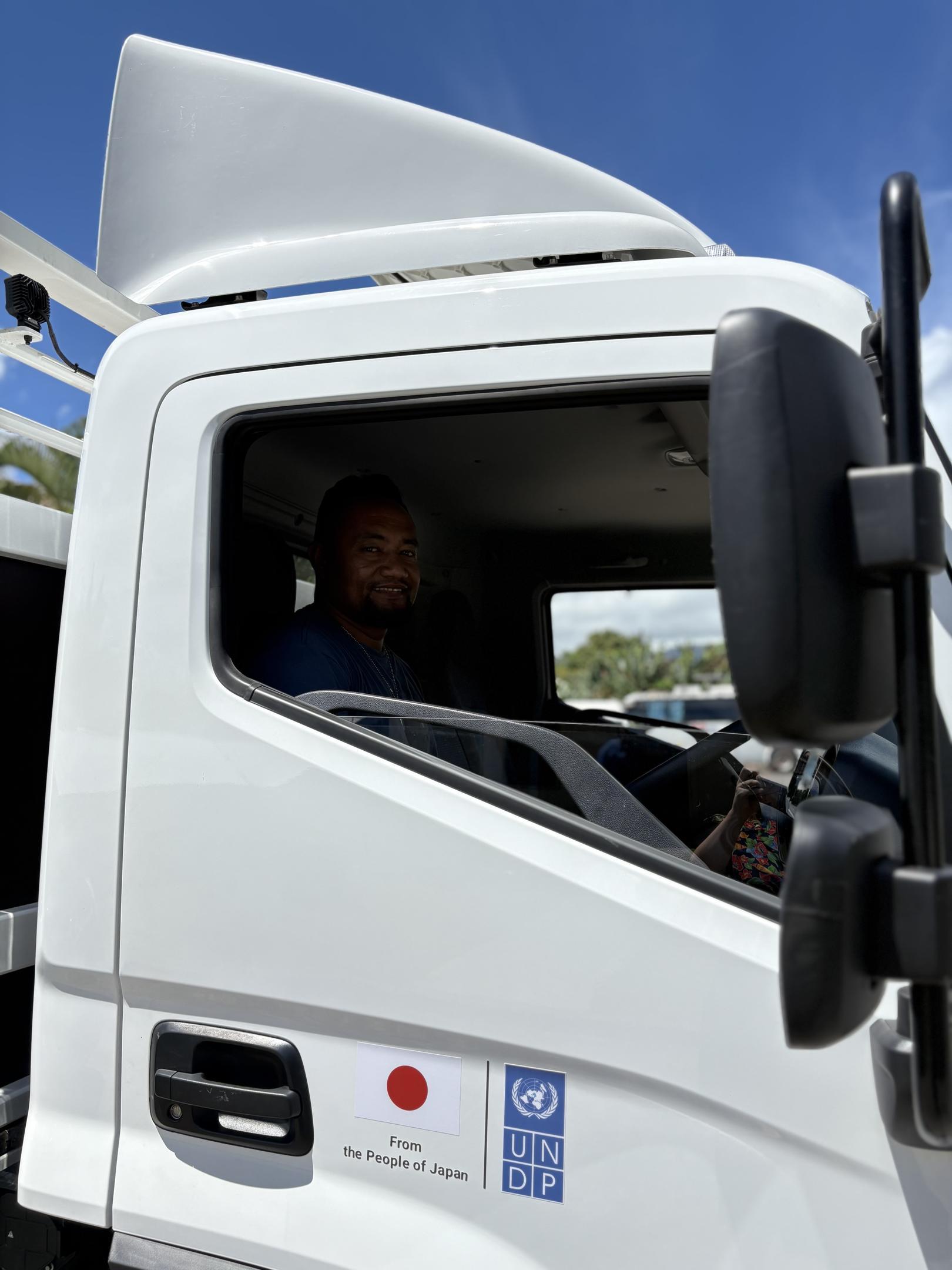 Photograph of a driver in the cab of a white truck, window open, blue sky behind.