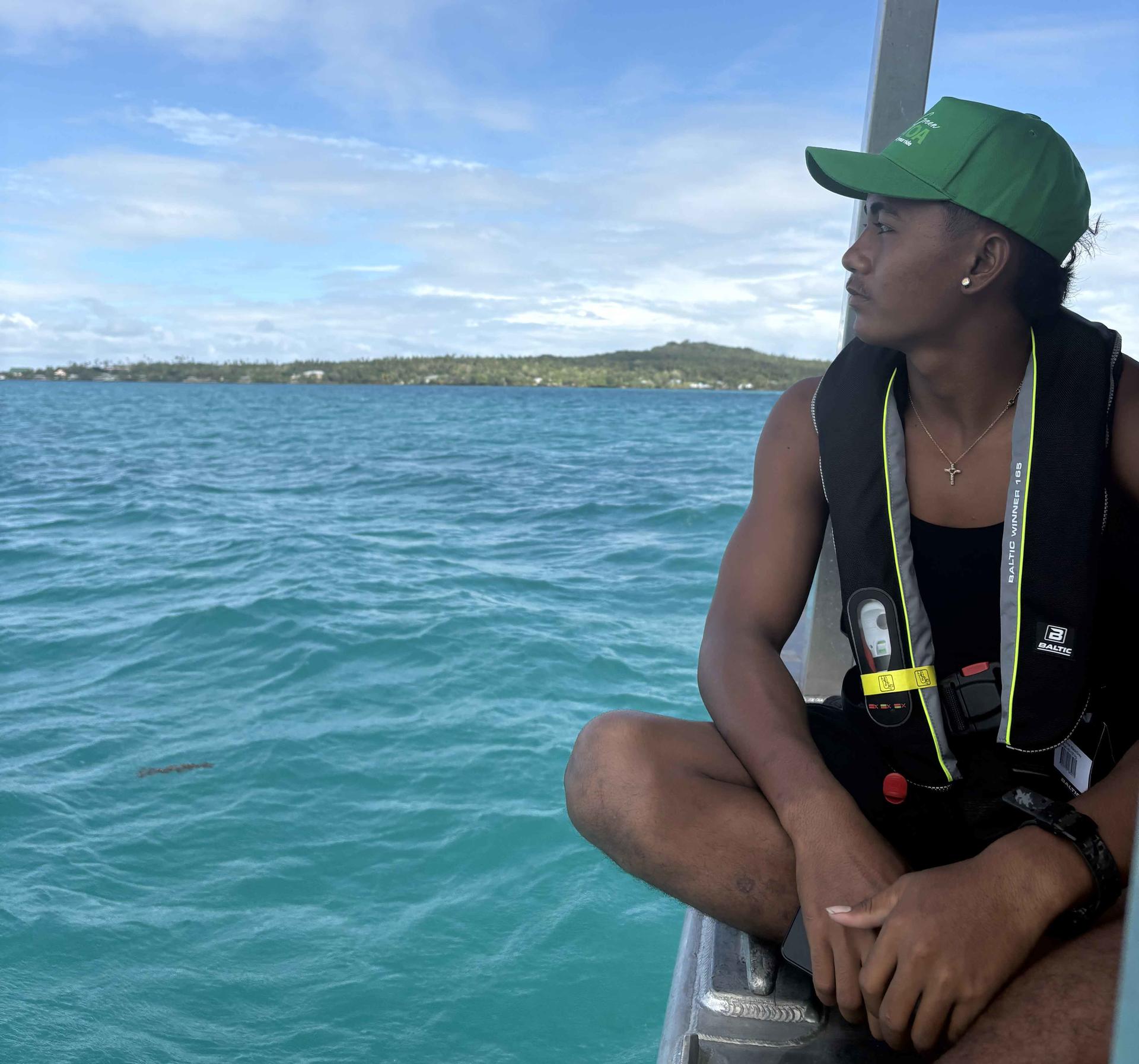 Person in a green cap and life jacket sits on a boat railing above turquoise water.