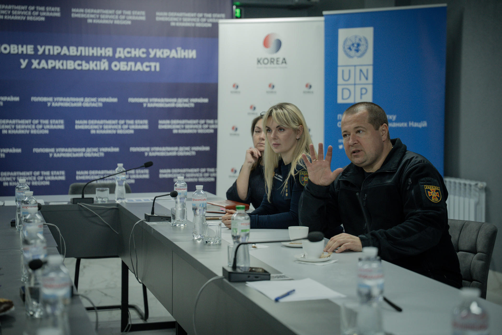 A meeting of the State Emergency Service of Ukraine in Kharkiv Oblast, where a man from the SES speaks and gestures with his hand as he sits at a conference table. Two women colleagues sit beside him and listen attentively. UNDP and Republic of Korea banners stand behind the speakers, with microphones, documents, water bottles, and cups on the table.