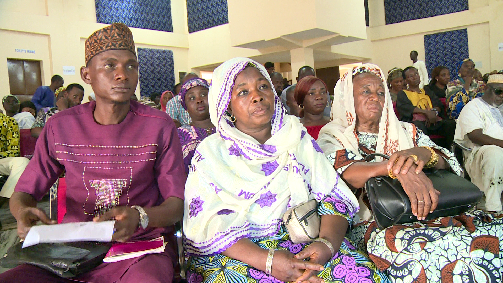 Audience of diverse people in a hall, wearing vibrant traditional clothing at a community gathering.