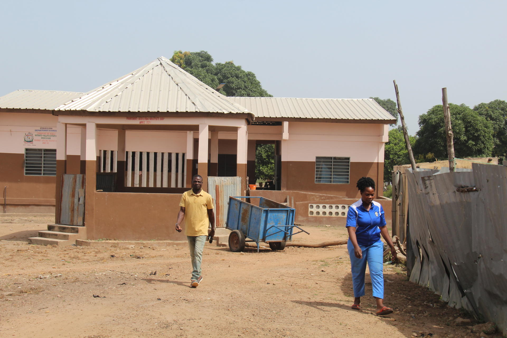 Two men stand near a tan, single-story building with a blue cart on a dusty yard.