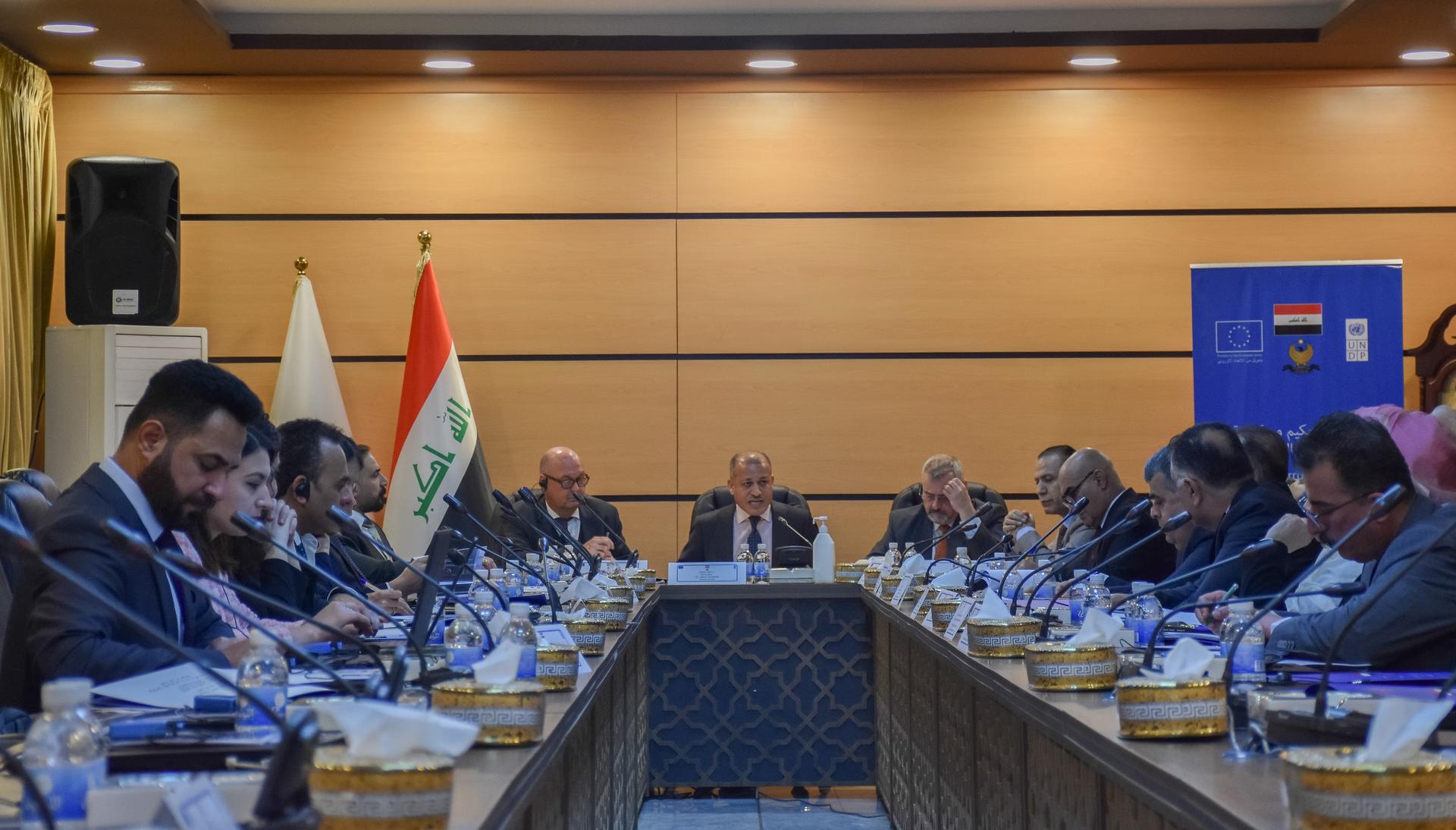 Formal meeting in a wood-paneled room with flags, participants seated around a long conference table.