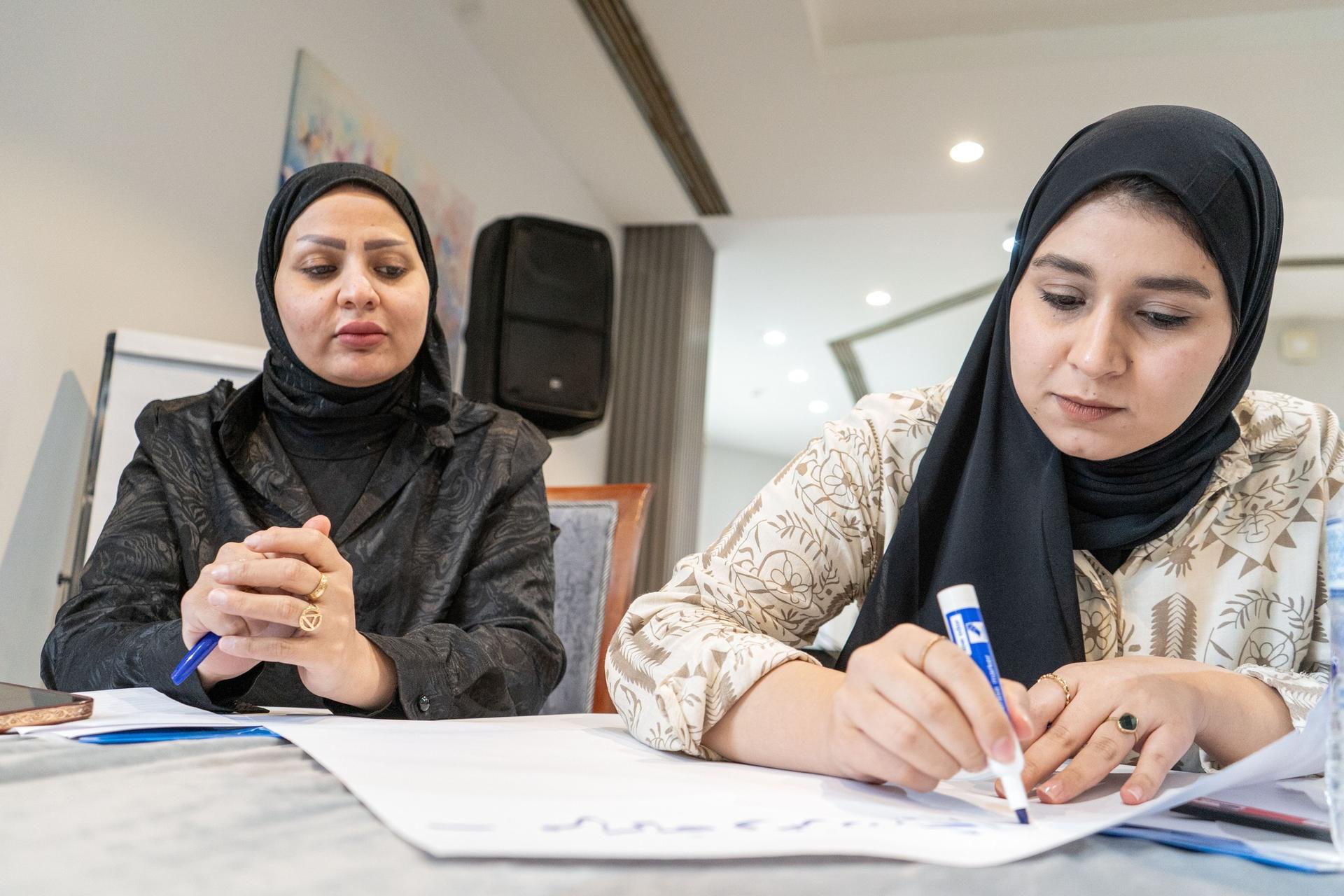 Two women sit at a table with documents; one writes with a blue pen.