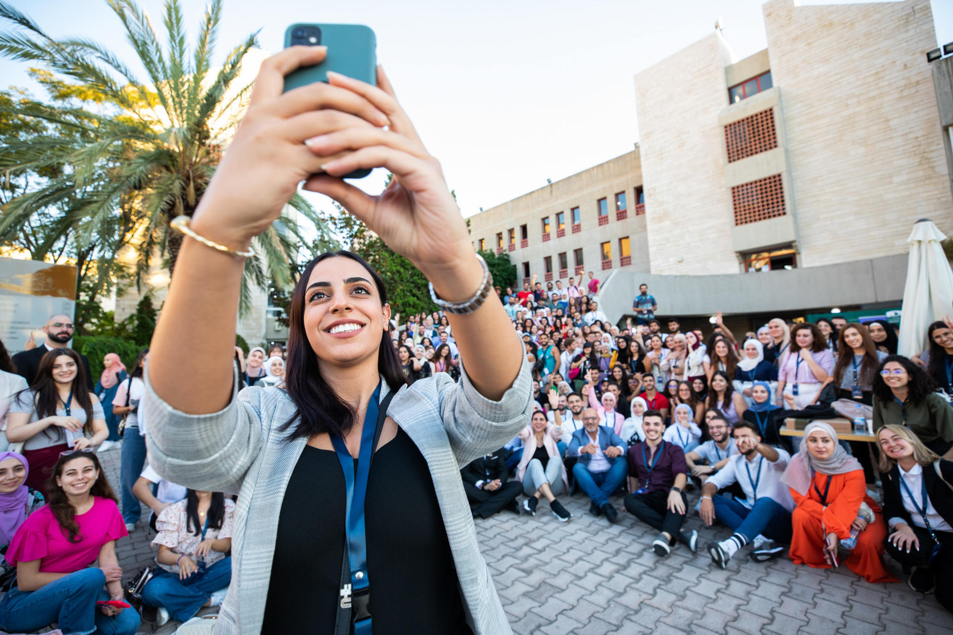 Person in foreground holds a blue smartphone up; crowd with palm trees and buildings behind.
