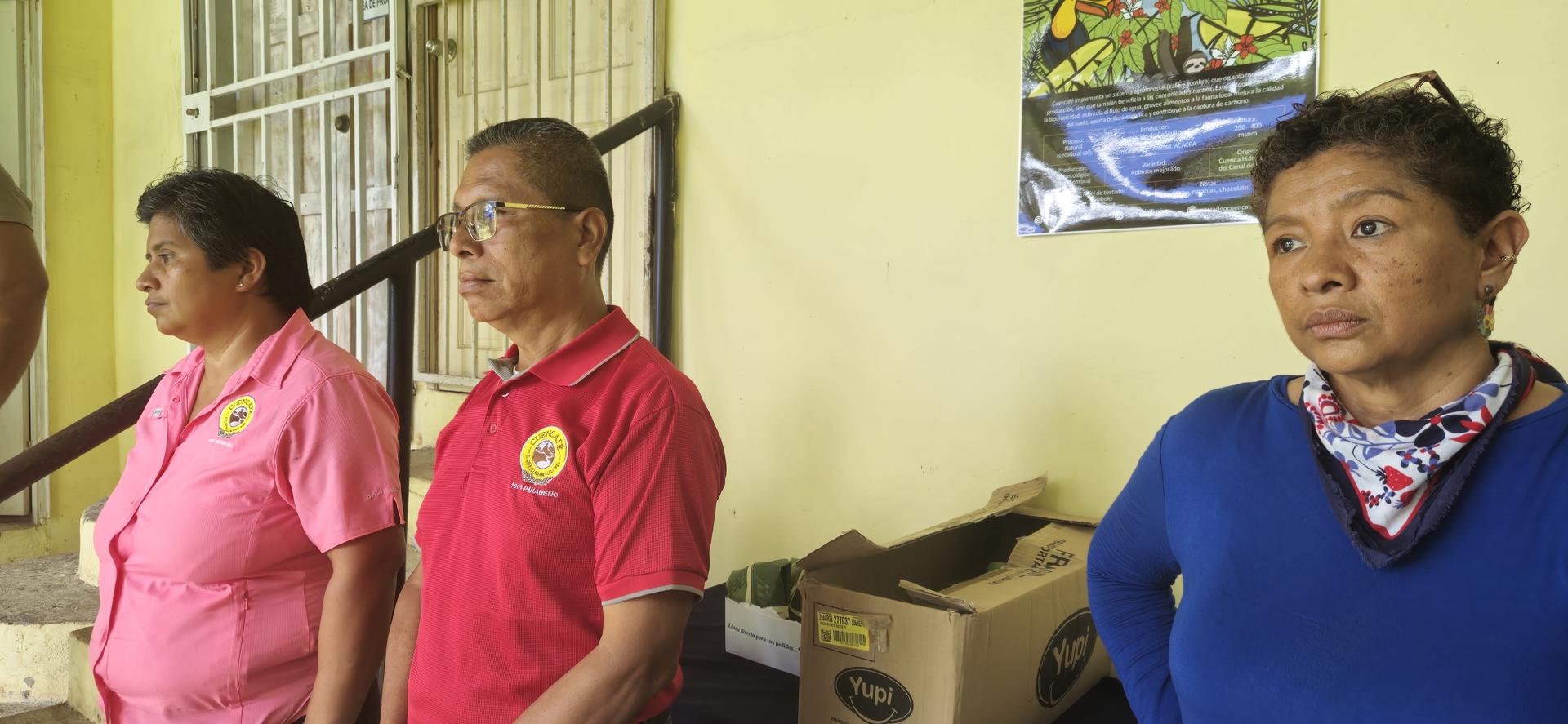Cuatro adultos están de pie en interiores junto a una pared con afiches; uno lleva una blusa azul y una bufanda