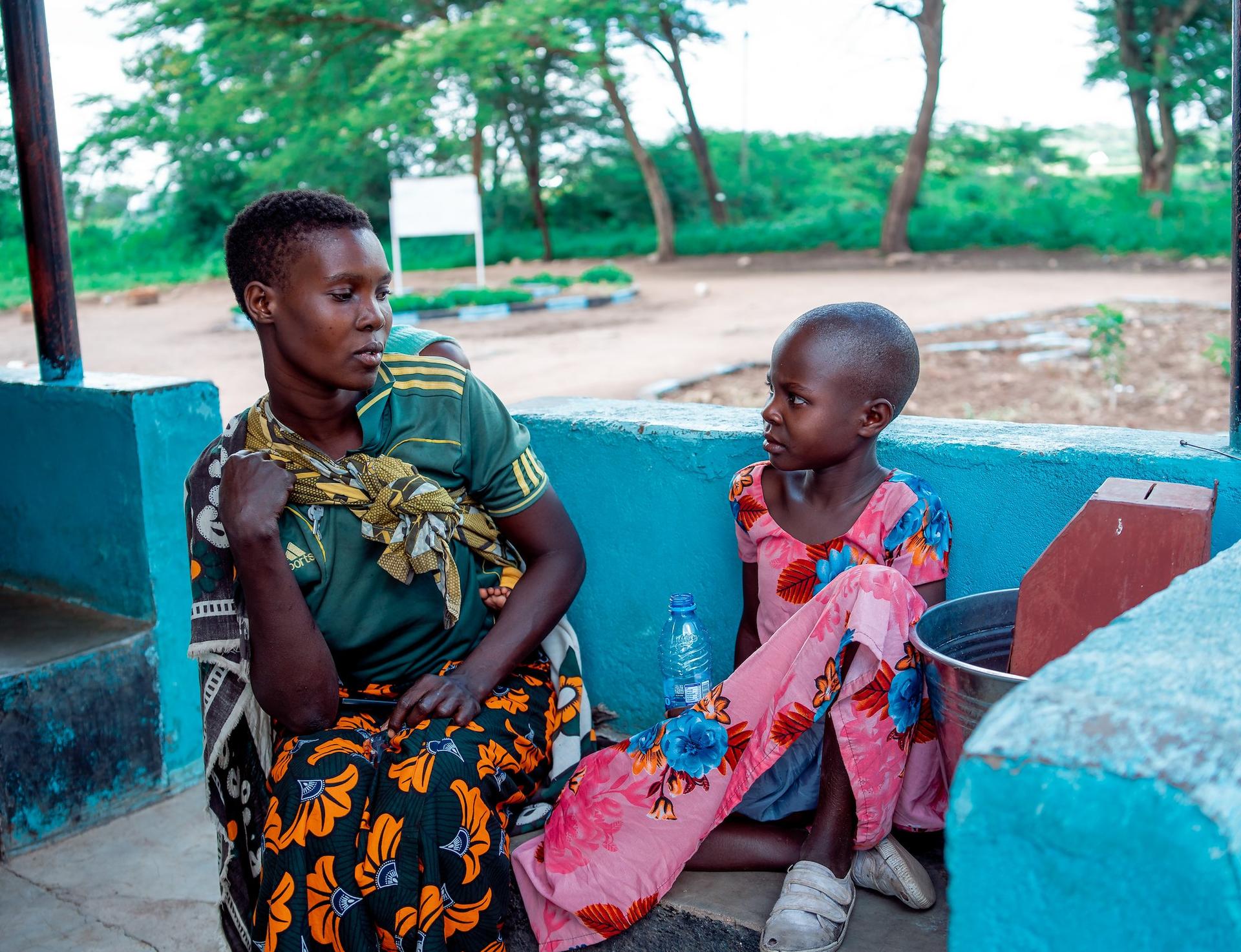 Two young children sit on a bench beneath a blue outdoor shelter, with green trees in the background.
