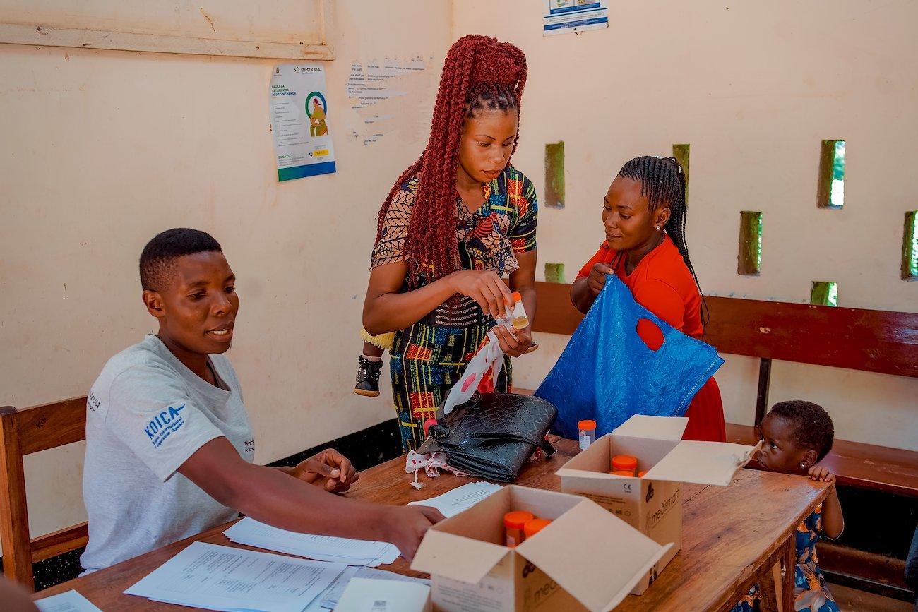 Teacher assists two students at a desk with papers in a small classroom.