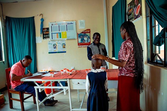 Photograph of a classroom craft session; a girl in blue stands while others work with orange paper.