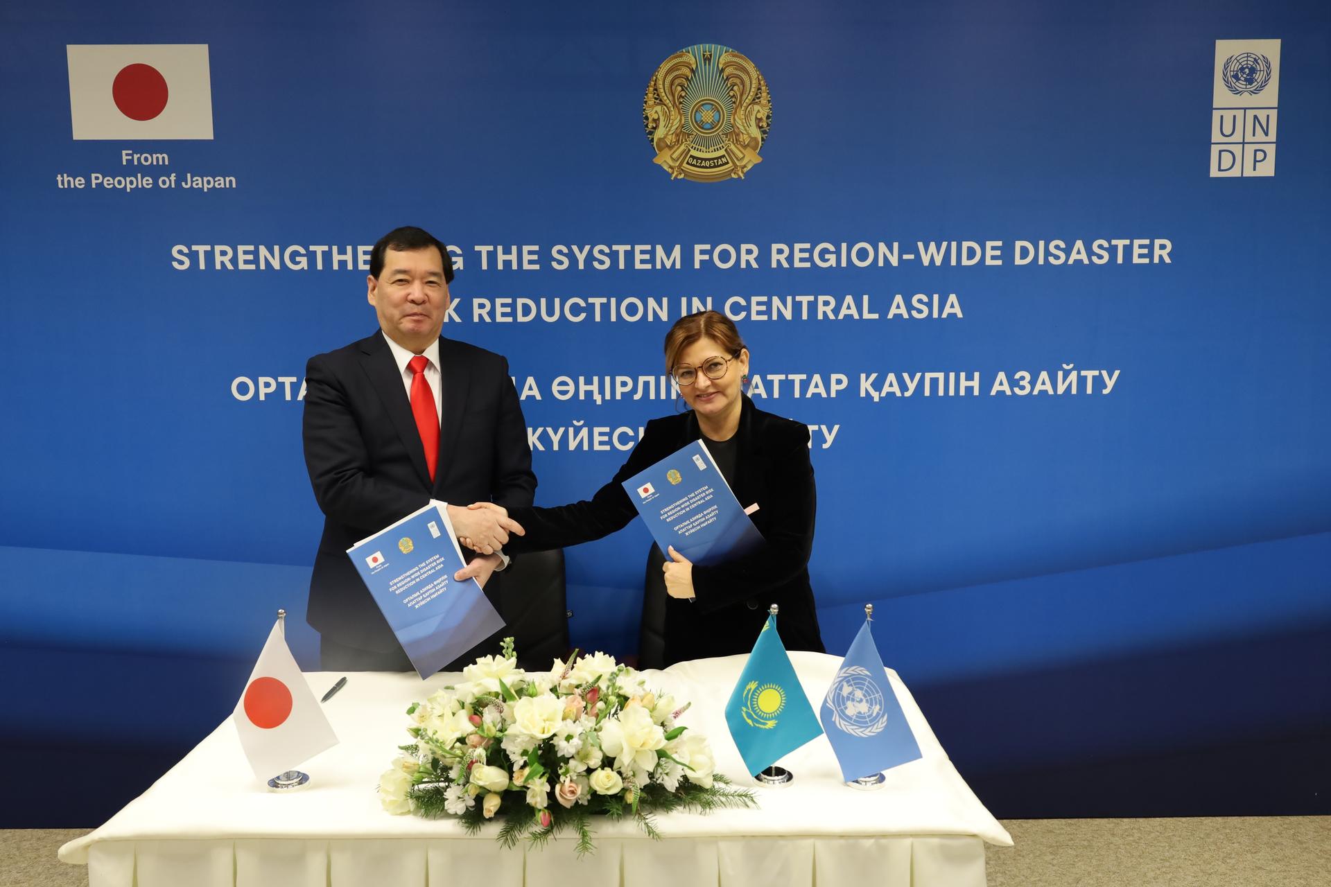 Two men in suits sign a document during a ceremony, with the Japanese flag and another flag, blue backdrop.