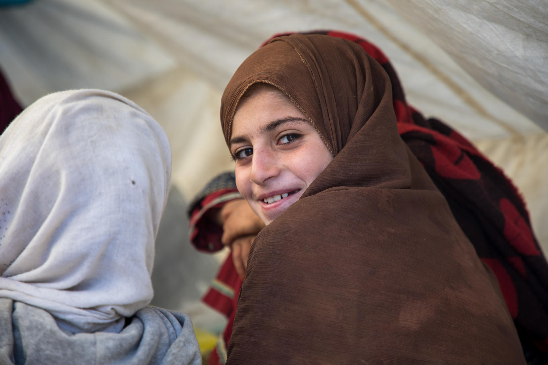 Two people in hijabs inside a tent, seen from behind; white and brown scarves.
