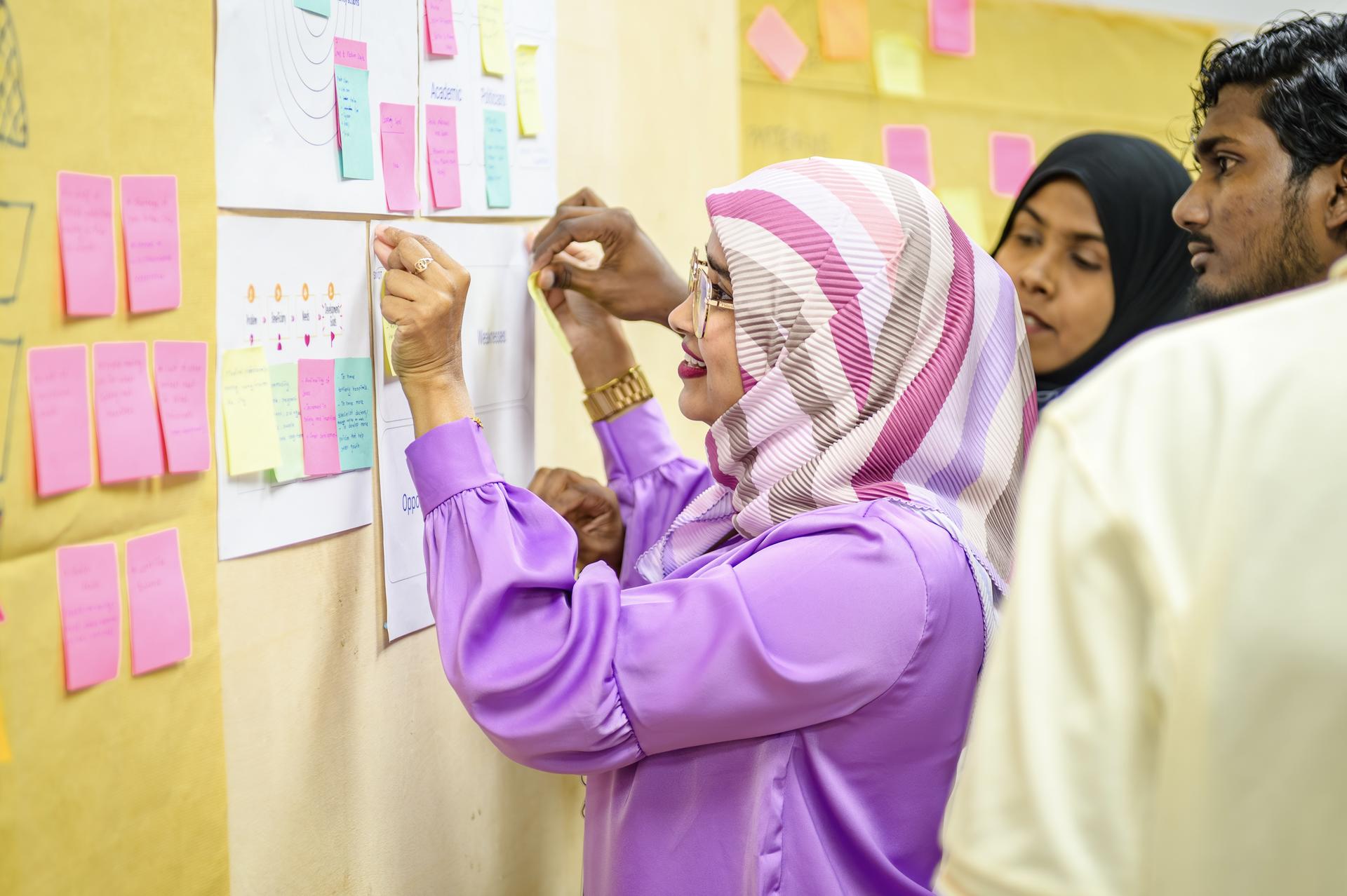 Woman in purple hijab sticks sticky notes onto a colorful wall.