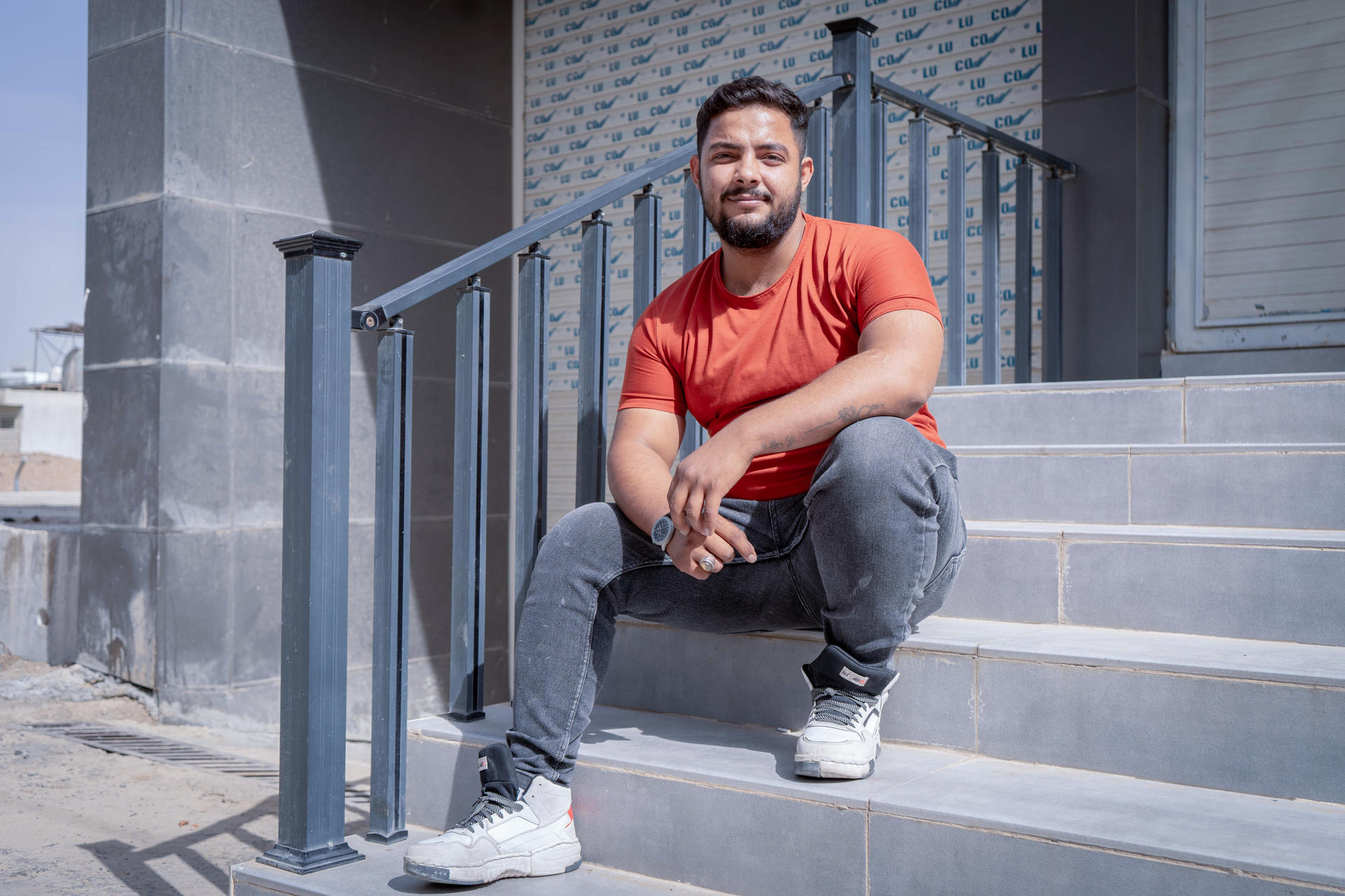 Bearded man sits on blue steps outside a building, wearing an orange shirt and gray pants, smiling.
