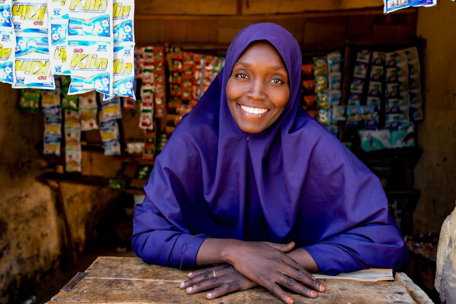 Person in a purple hijab sits behind a wooden counter in a shop, hands folded on the surface.