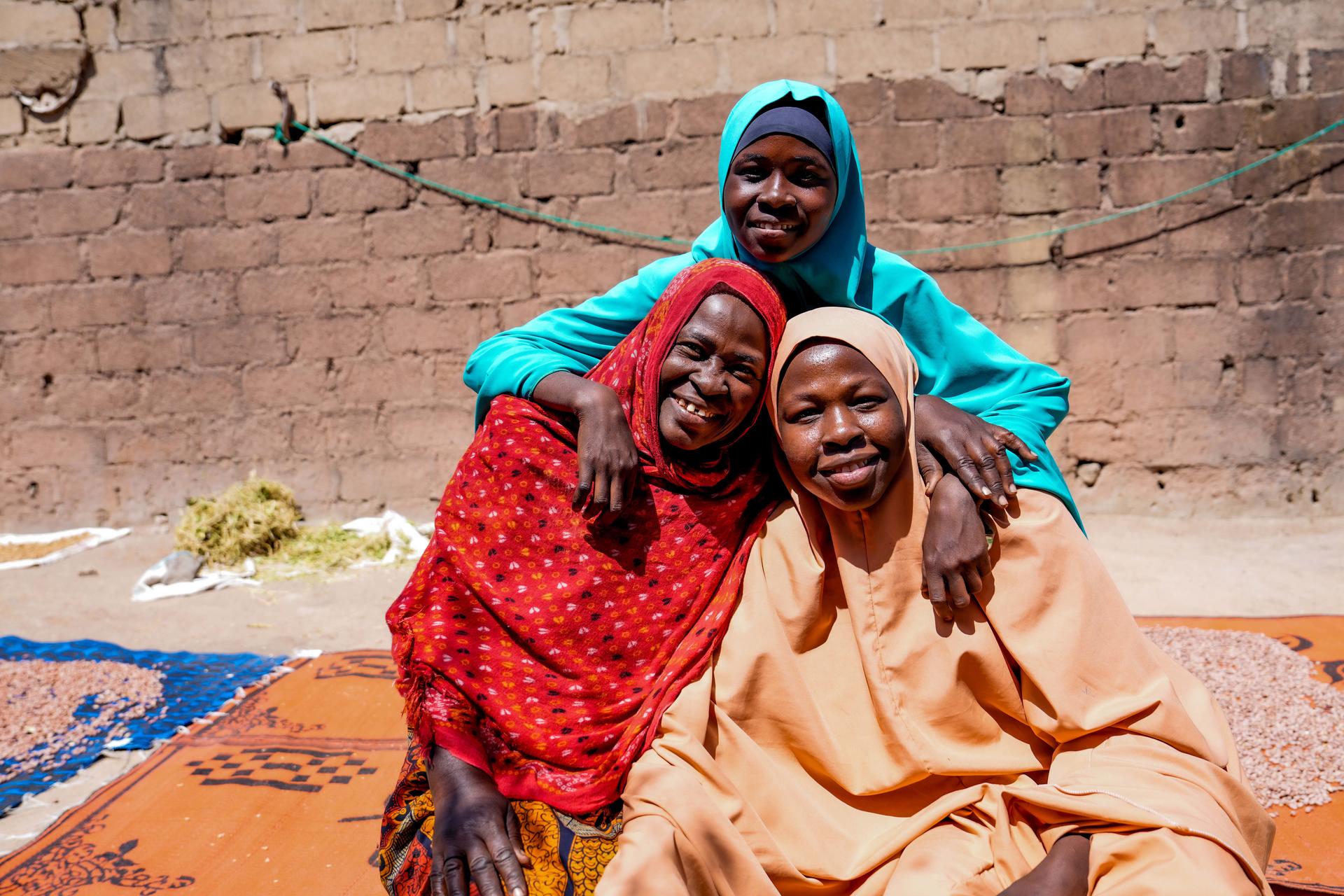 Three women in vibrant traditional attire pose on colorful mats outdoors, brick wall behind.