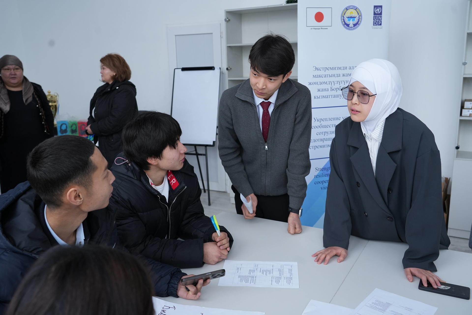 Photo of students around a table as a suited man talks to a woman in a hijab; papers spread out.