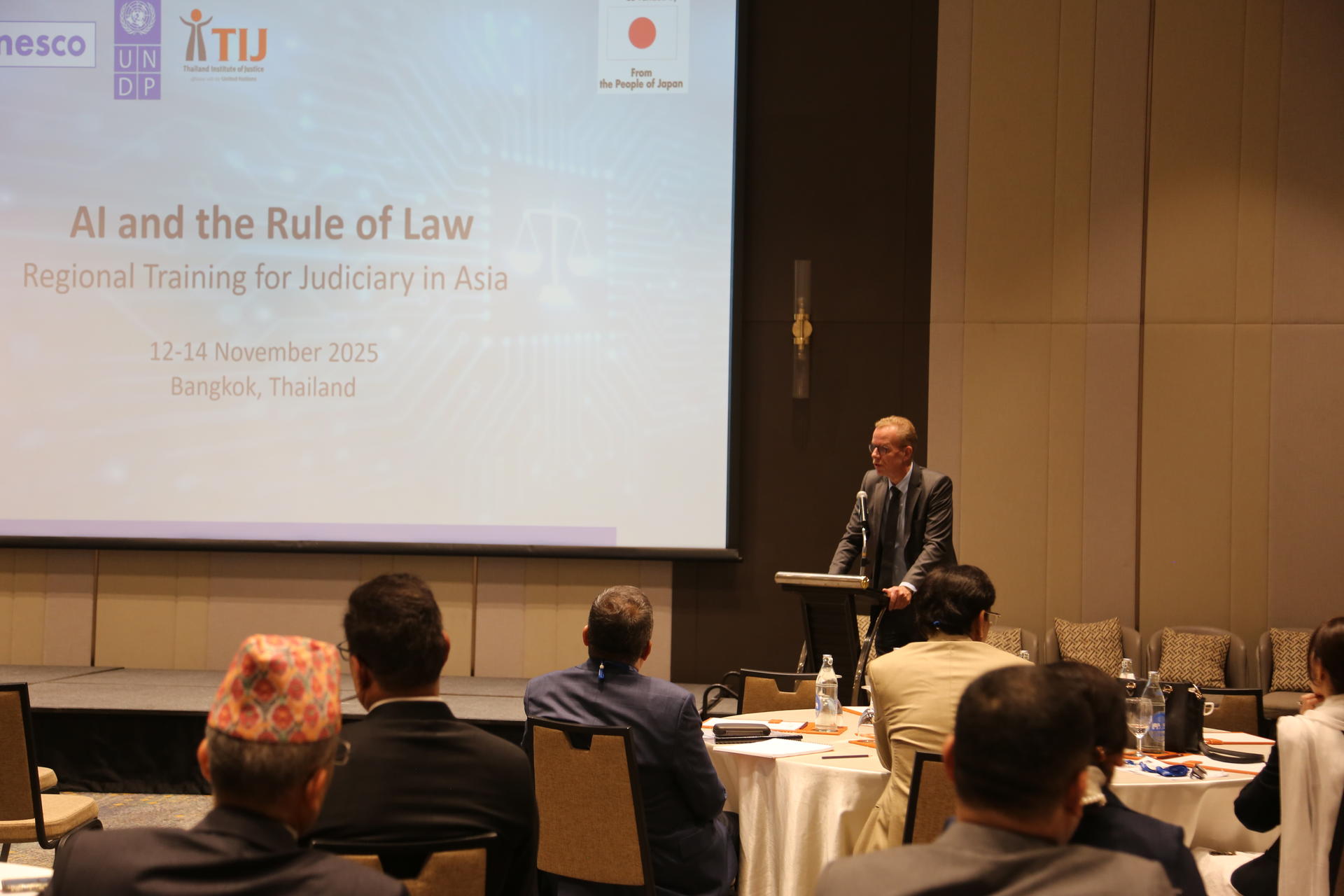 Conference room presentation: speaker at a podium presenting a slide titled All and the Rule of Law.