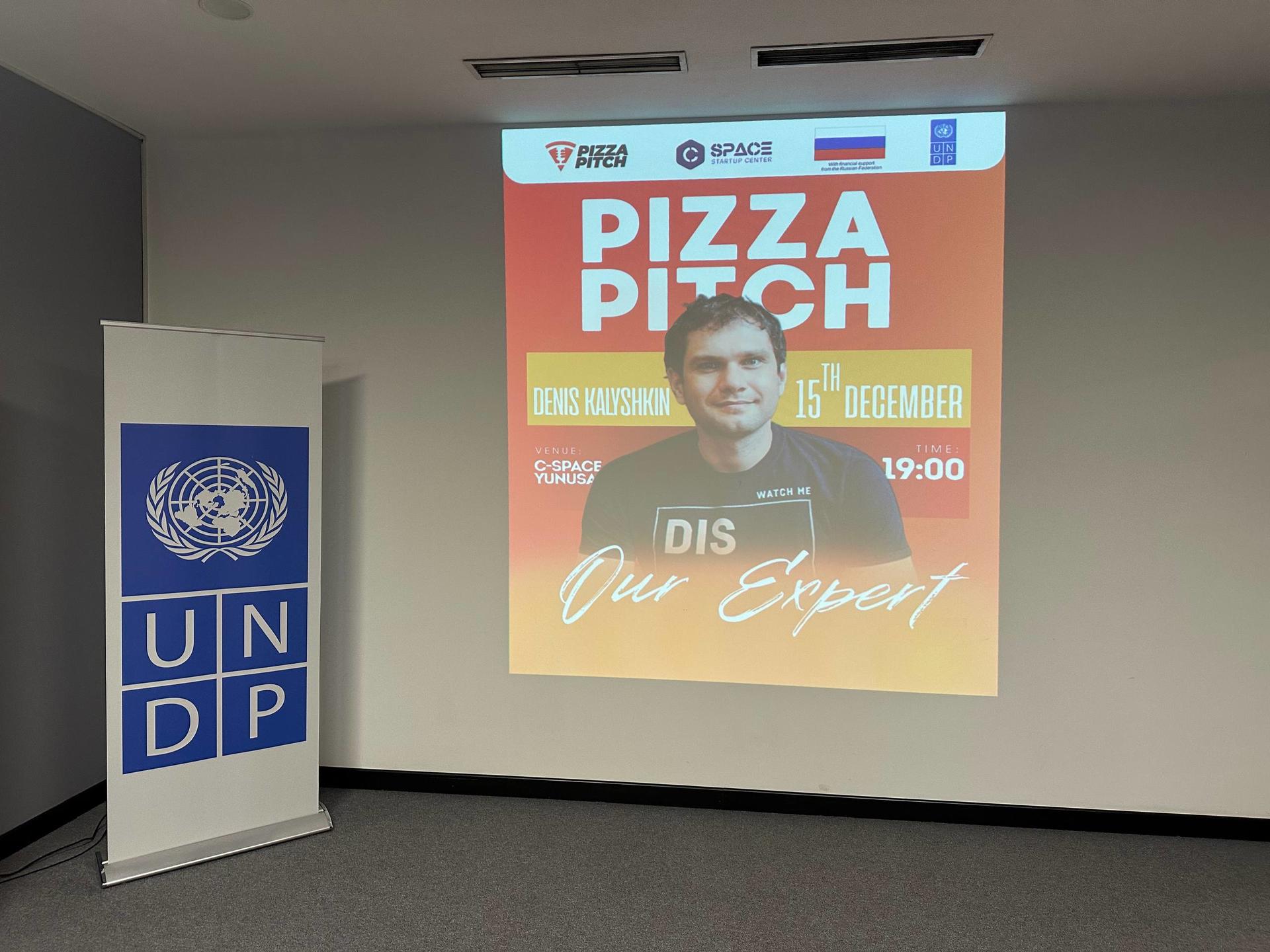 Conference room with an UNDP banner; projection displays a Pizza Night poster featuring a man.