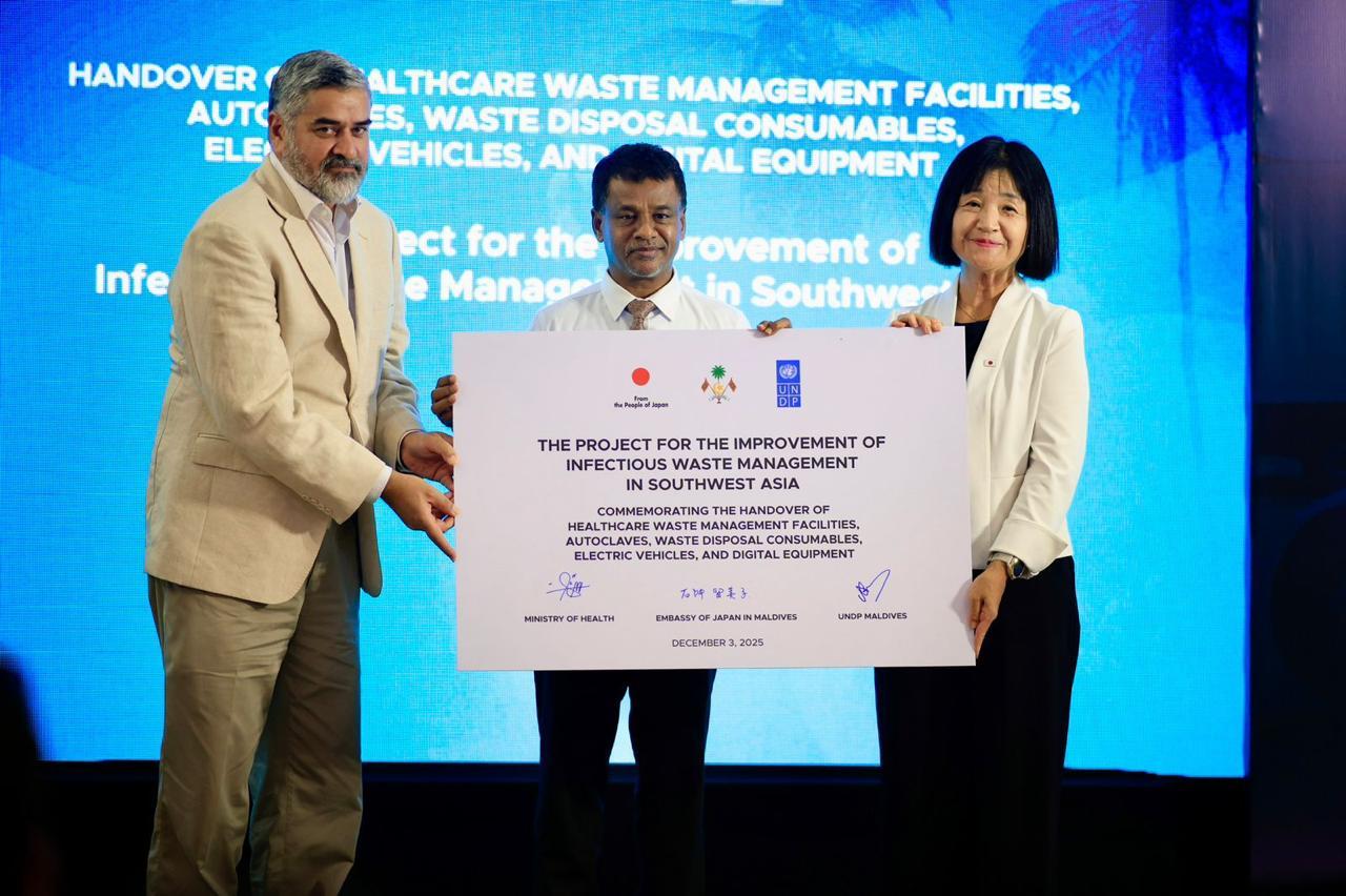 Three people on a stage presenting a large certificate against a blue backdrop.