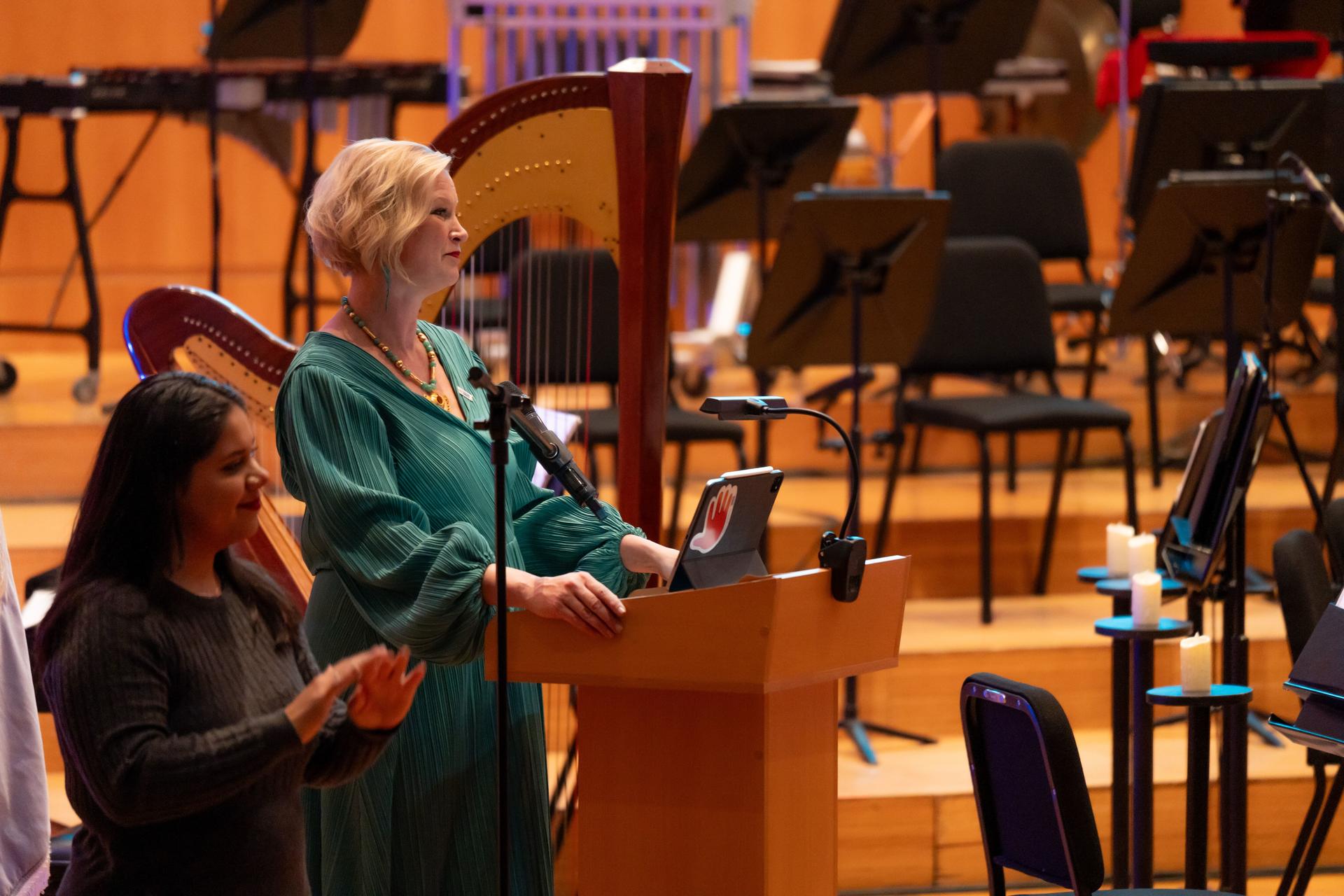 Woman in teal at podium speaking; sign language interpreter beside her; orchestra in background.