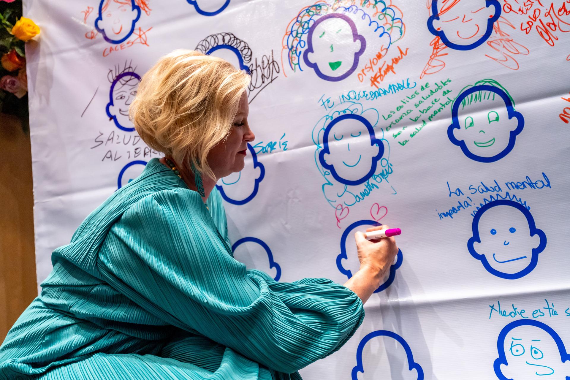 Woman in a teal jacket drawing with an orange marker on large banner of blue outlined faces.