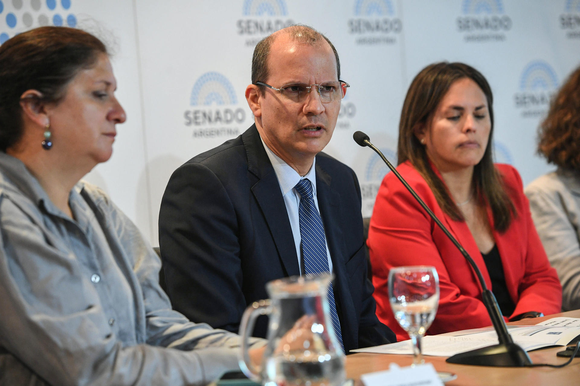Three attendees at a formal panel; left in denim jacket, middle in suit, right in red blazer.