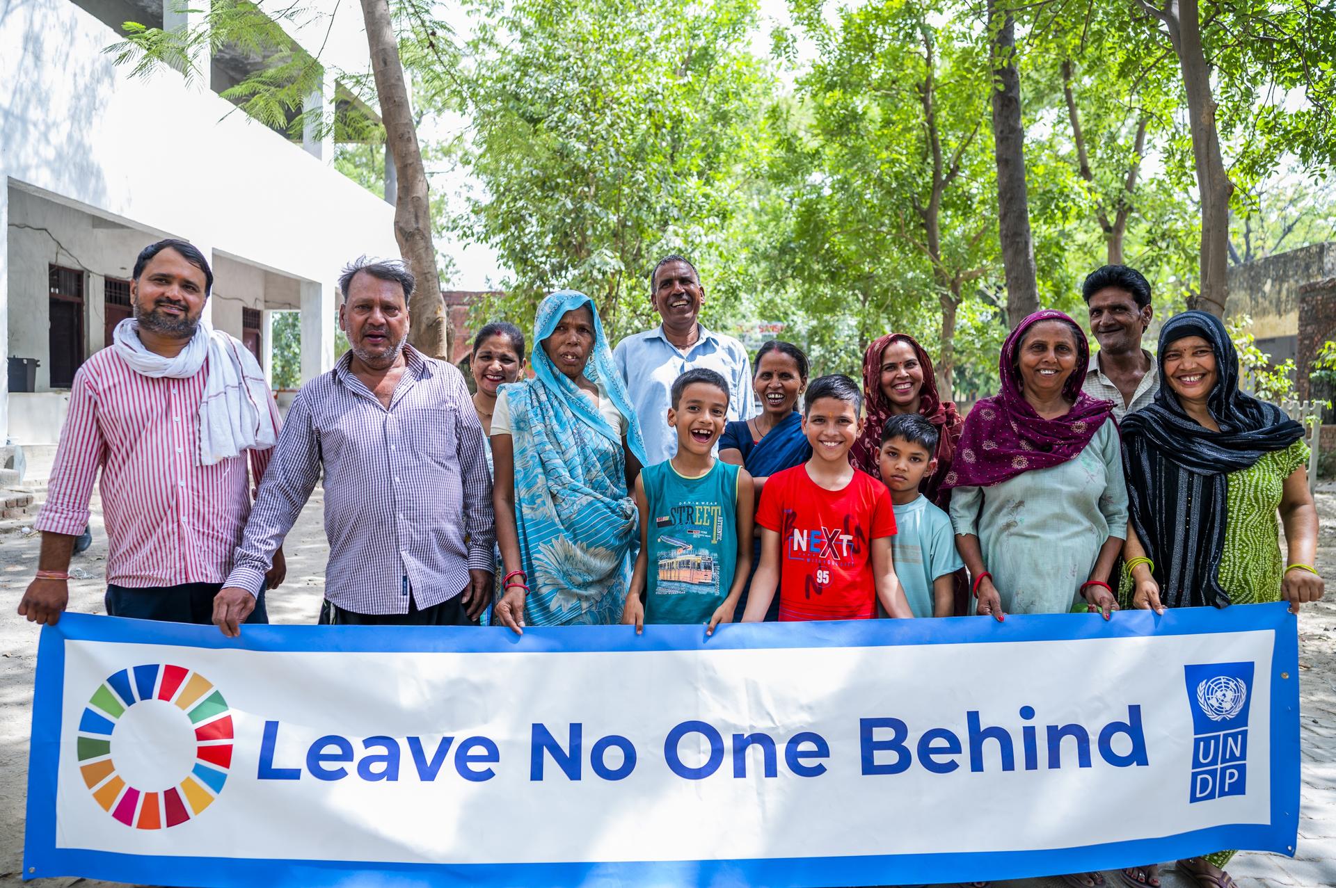 Diverse group outdoors smiling while holding a banner reading Leave No One Behind.