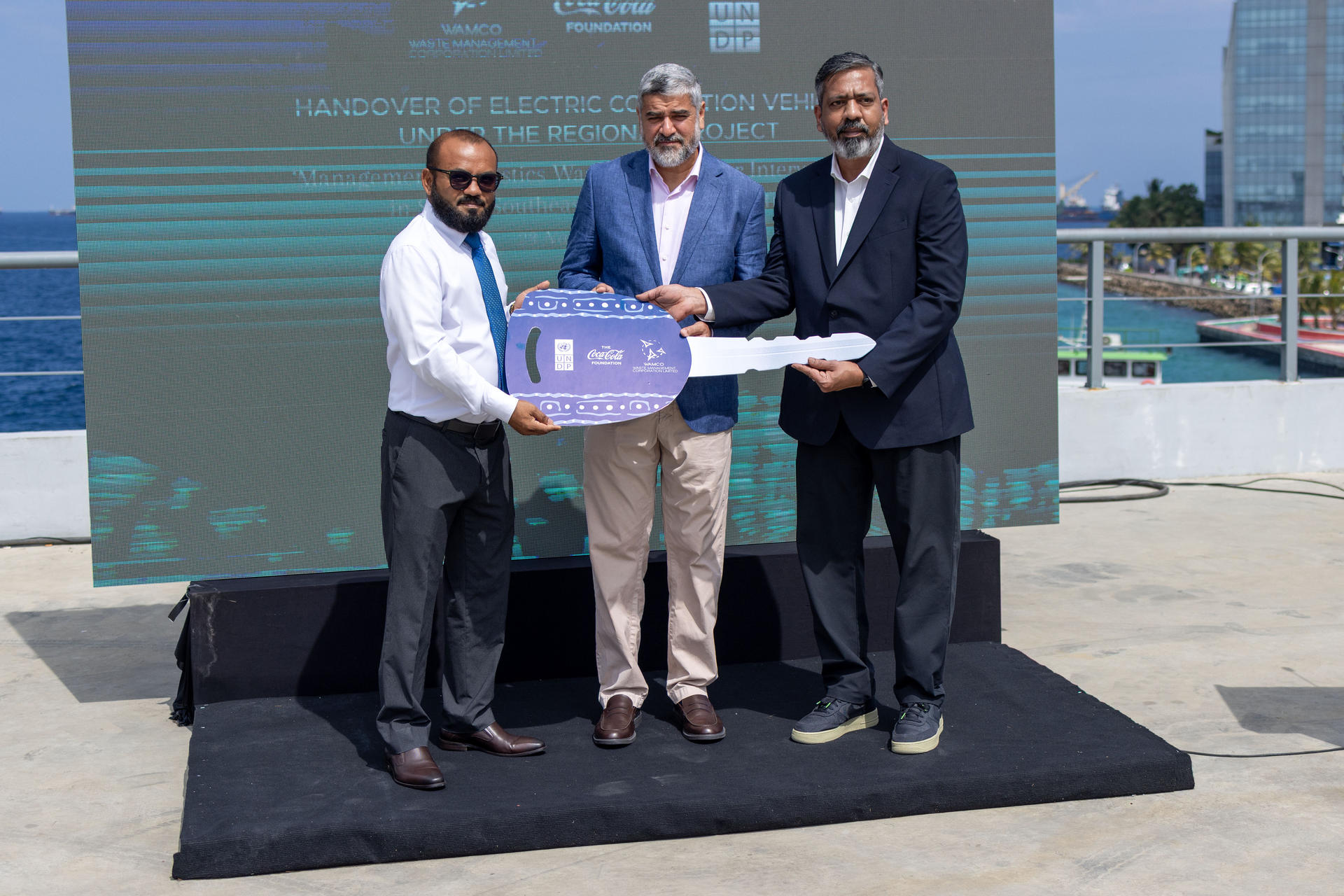 Four men in suits on an outdoor stage by the water, presenting a purple plaque.